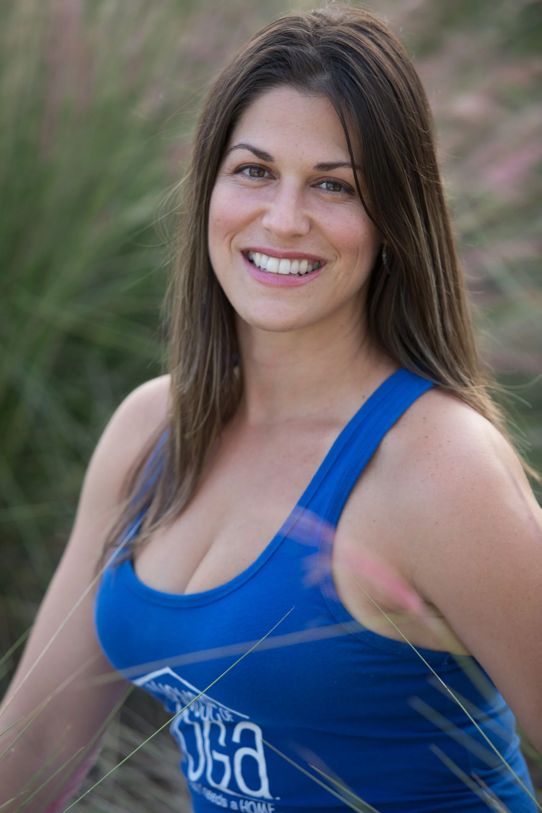 Close-up of a woman smiling outdoors, wearing a blue tank top, with long brown hair and brown eyes, surrounded by tall grass and greenery.