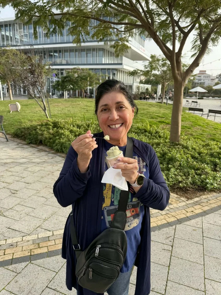 A smiling woman with dark hair in a ponytail eating ice cream outdoors in a park with greenery and trees, modern office building in the background, wearing a blue jacket, a graphic t-shirt, a watch, and a black crossbody bag.