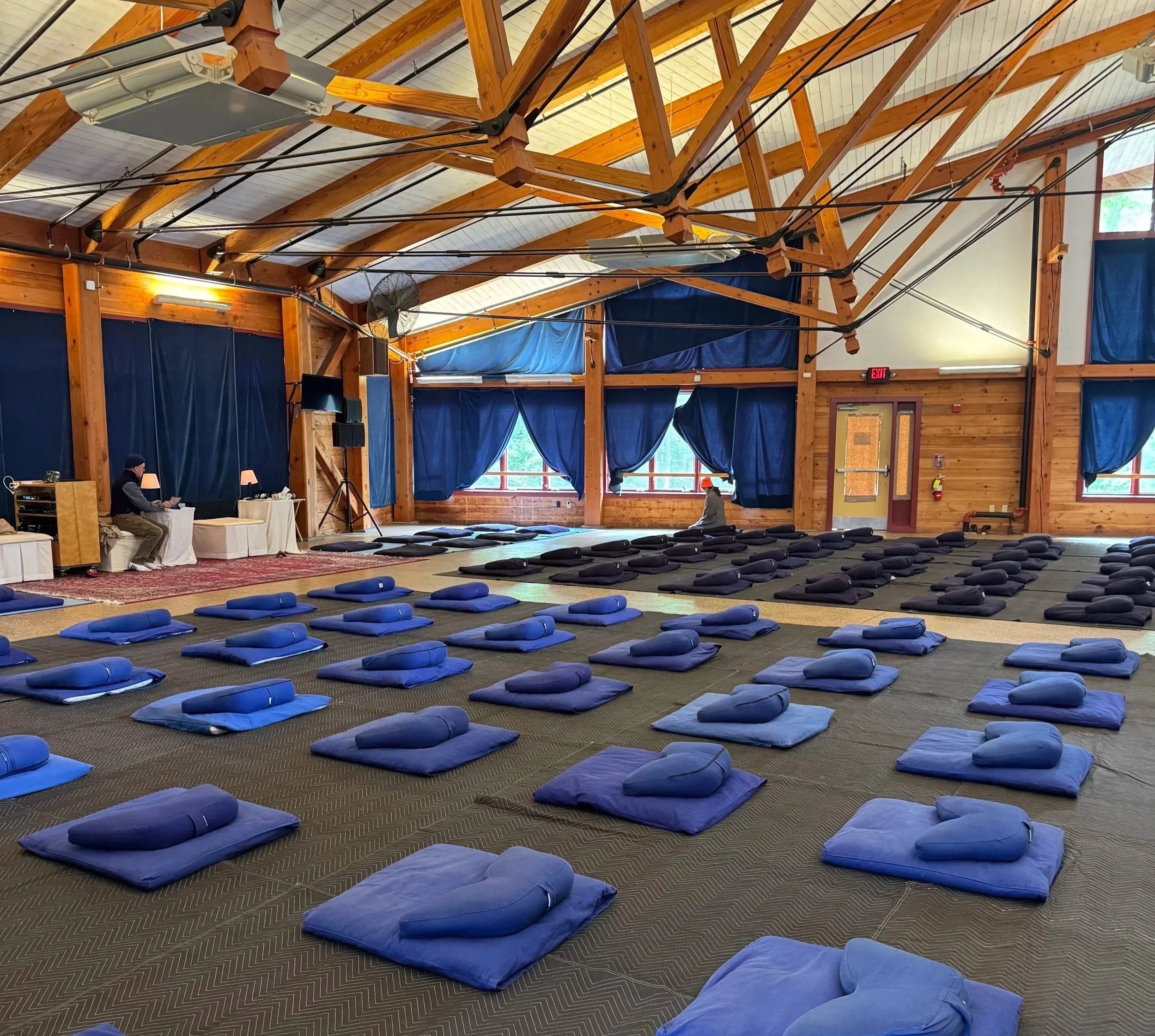 An indoor space set up for a meditation or yoga session with rows of blue cushions and pillows arranged on the carpeted floor, and a small stage area at the front with two people preparing for the session.