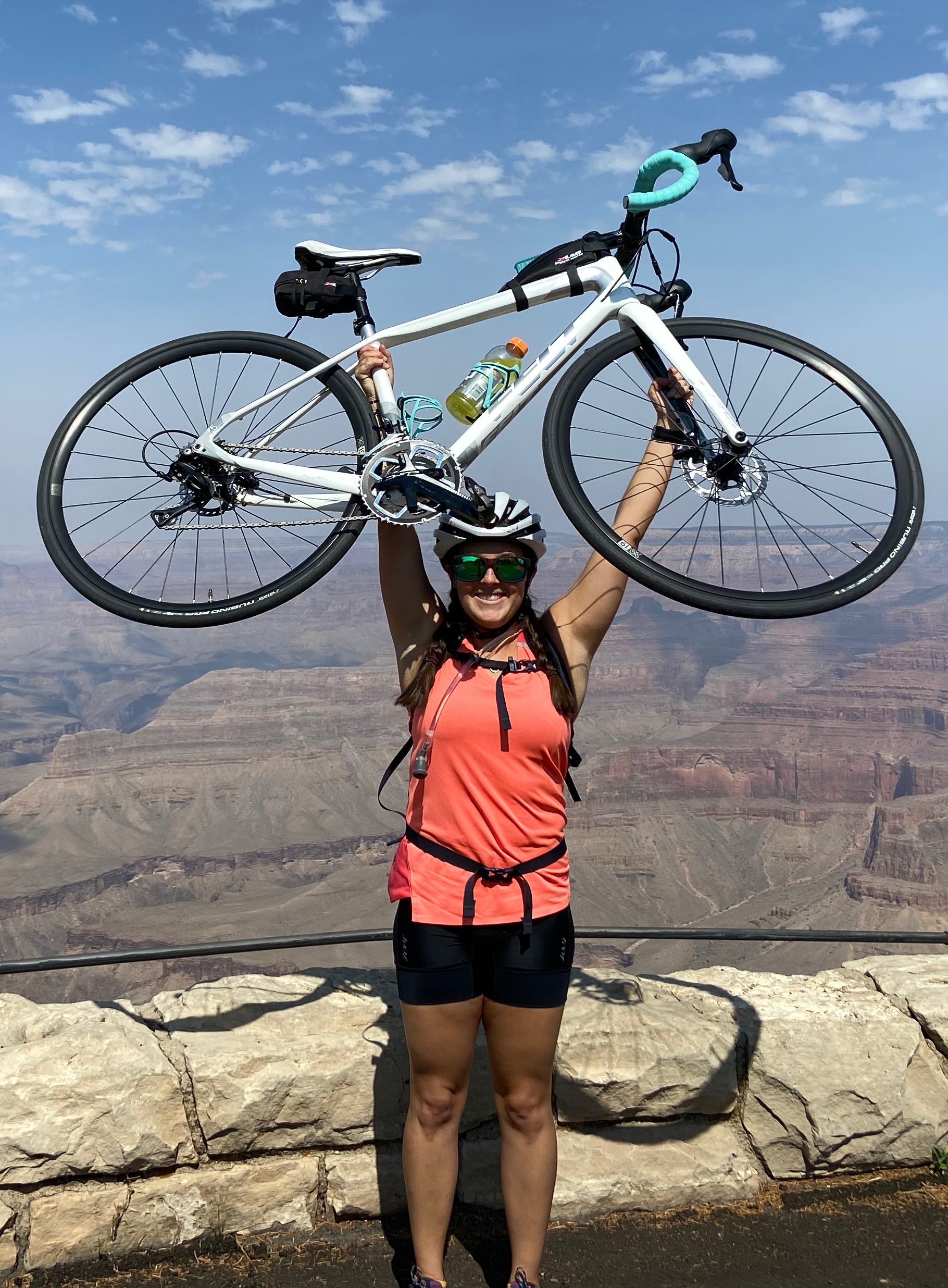 Woman in cycling gear, wearing a helmet and sunglasses, holding a white bicycle above her head at the Grand Canyon with a clear blue sky and canyon landscape in the background.