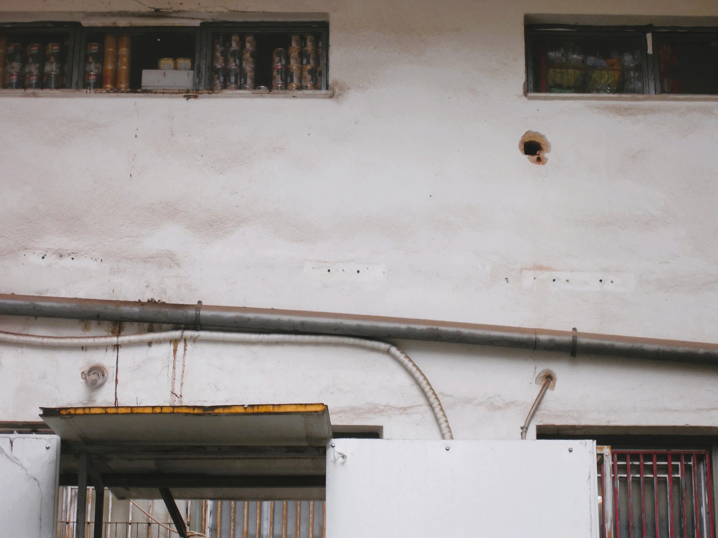 Close-up of a white building wall with small windows at the top, some pipes, and a small circular hole.