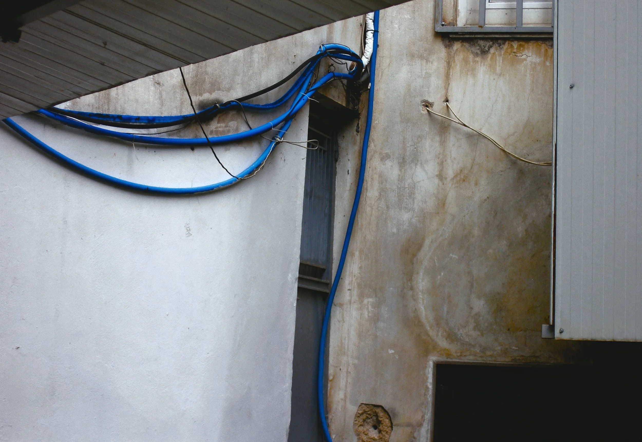 Exterior wall with multiple blue electric cables and wires running along it, some tied together and hanging loosely, with a metal security grate window and a patch of damaged wall.