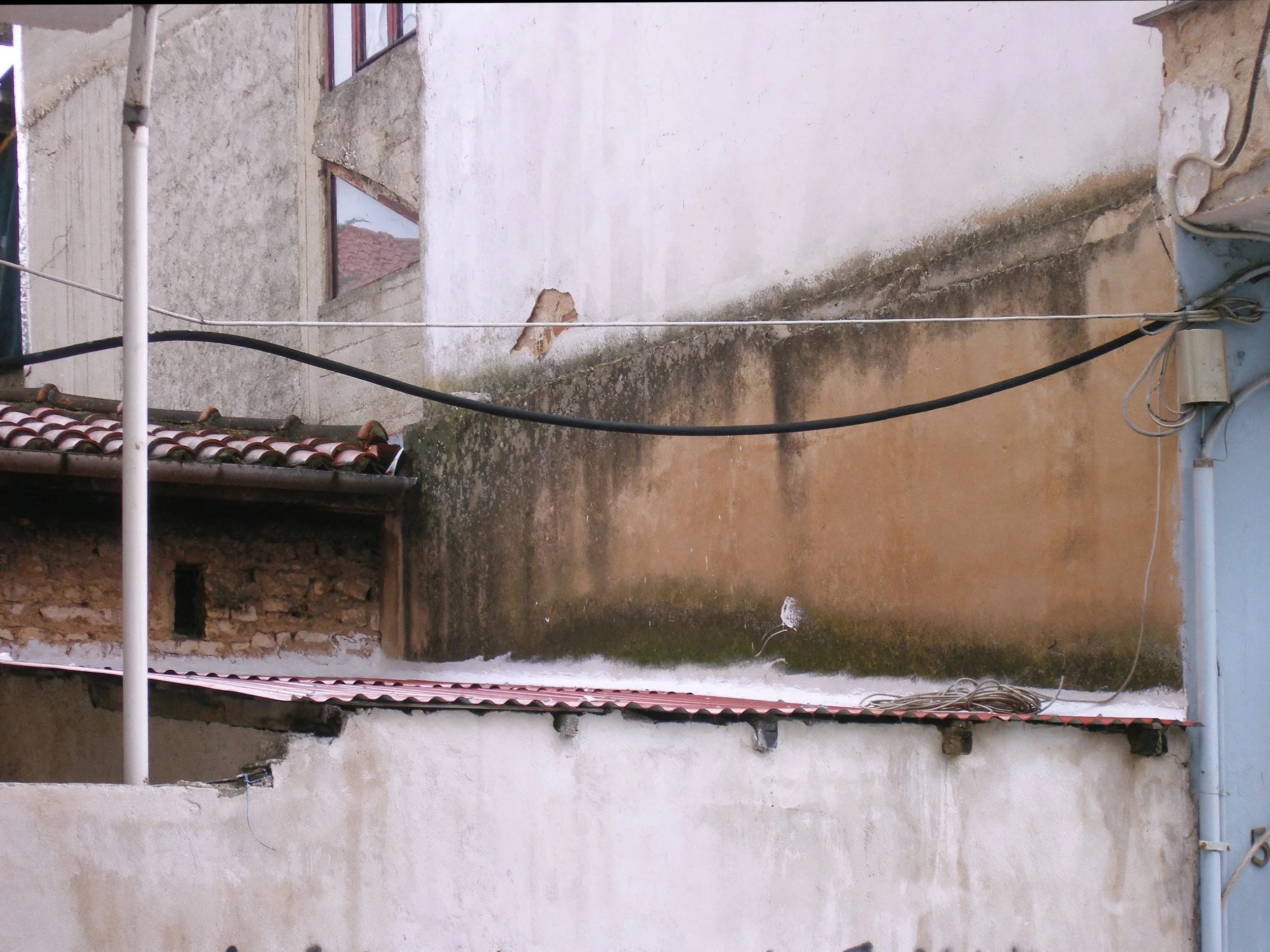 Side of a building with weathered walls, black and white electrical wires, and a small tiled roof structure.
