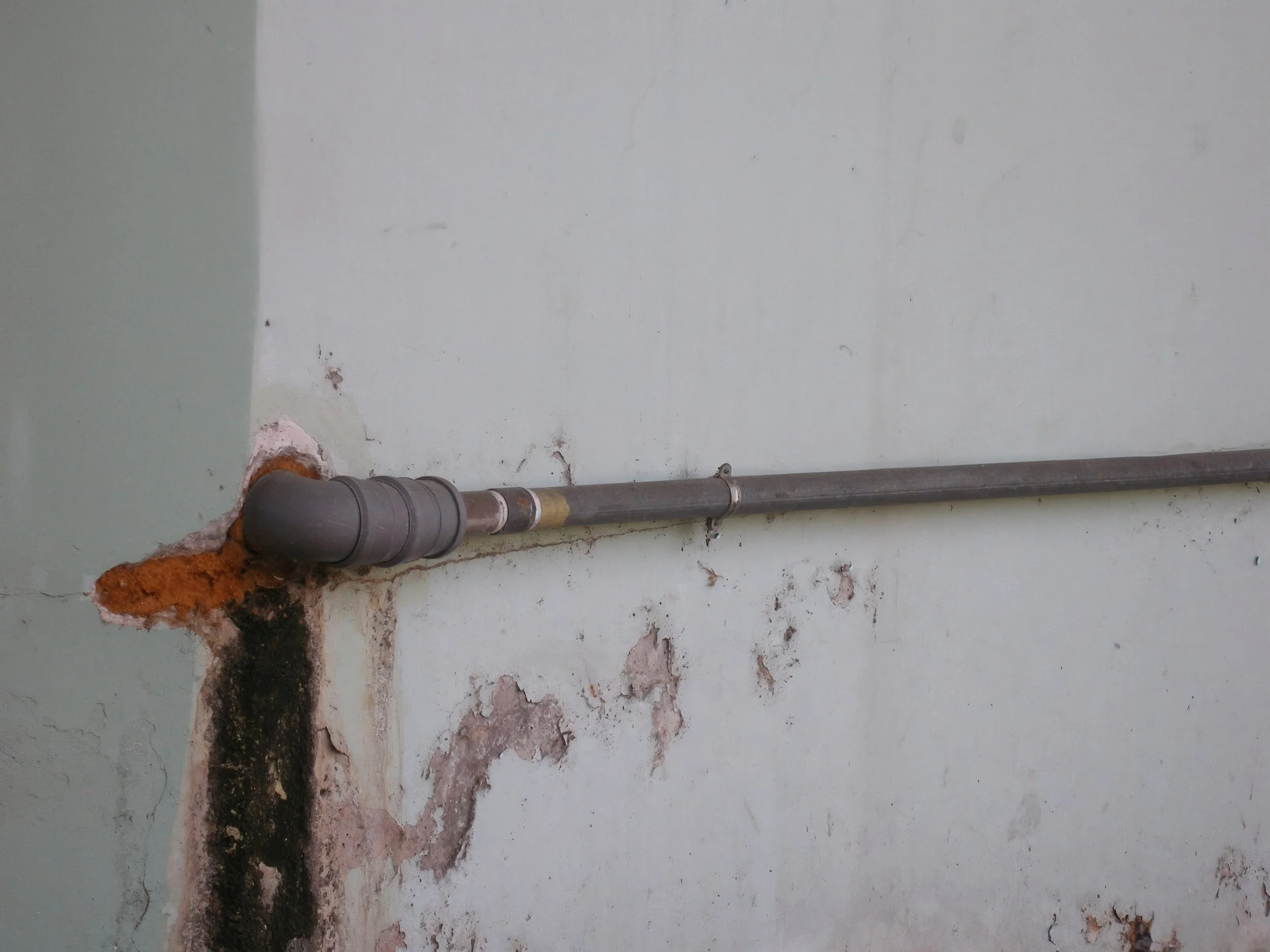 A metal pipe running horizontally along a wall, with visible stains and deterioration around the area where the pipe enters the wall.
