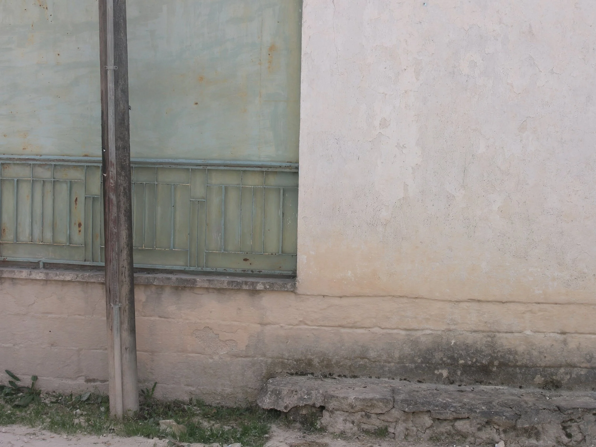 A weathered wooden utility pole in front of an old, textured wall with a window frame and a metal grate, on a patch of grass and dirt.