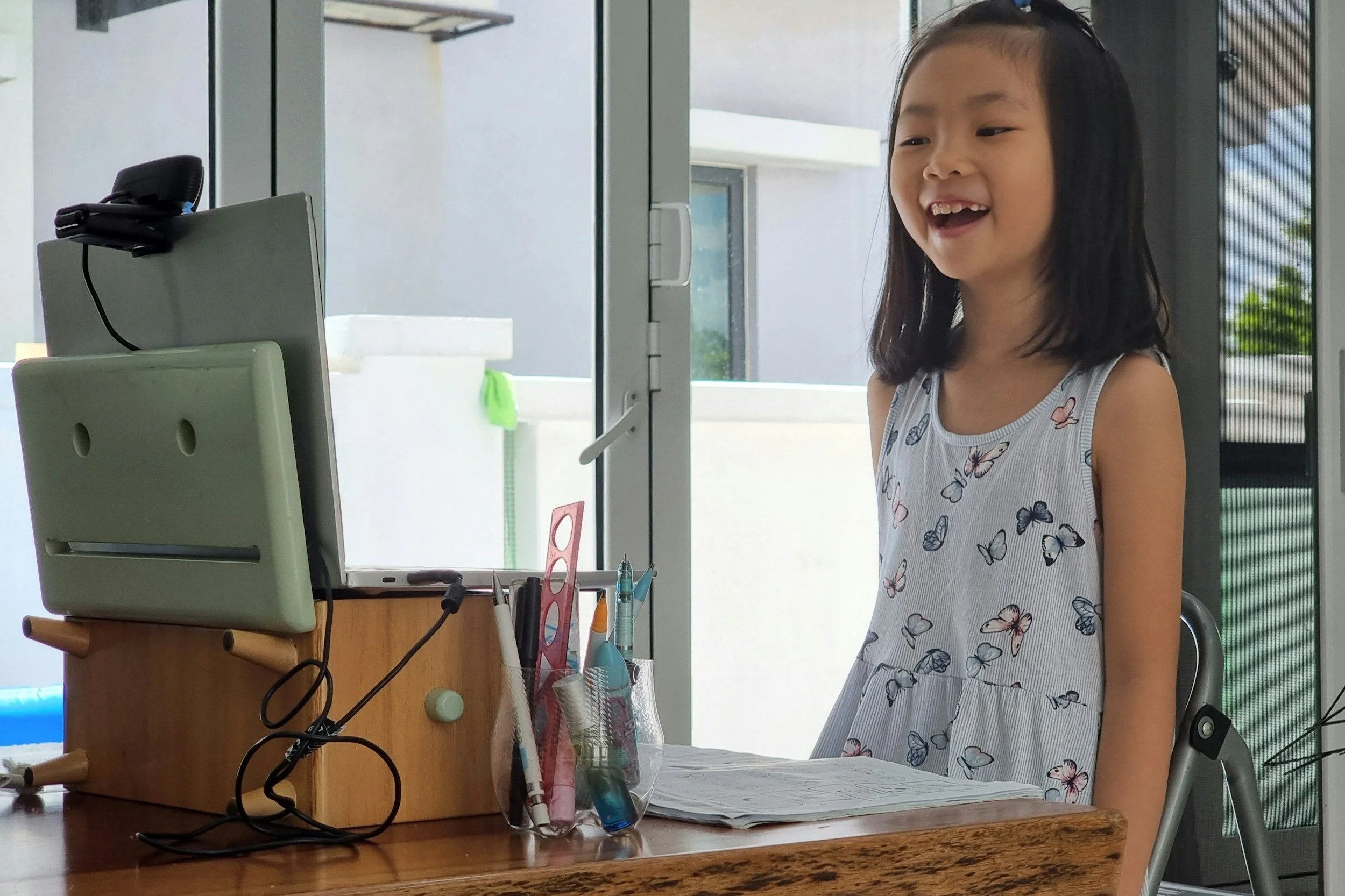 A young girl with long black hair and a butterfly-patterned sleeveless dress is smiling and looking at a computer monitor on a desk. The desk has various stationery items, including pens, a ruler, and paper. The background shows a glass door and an outdoor area with a bathtub and greenery.