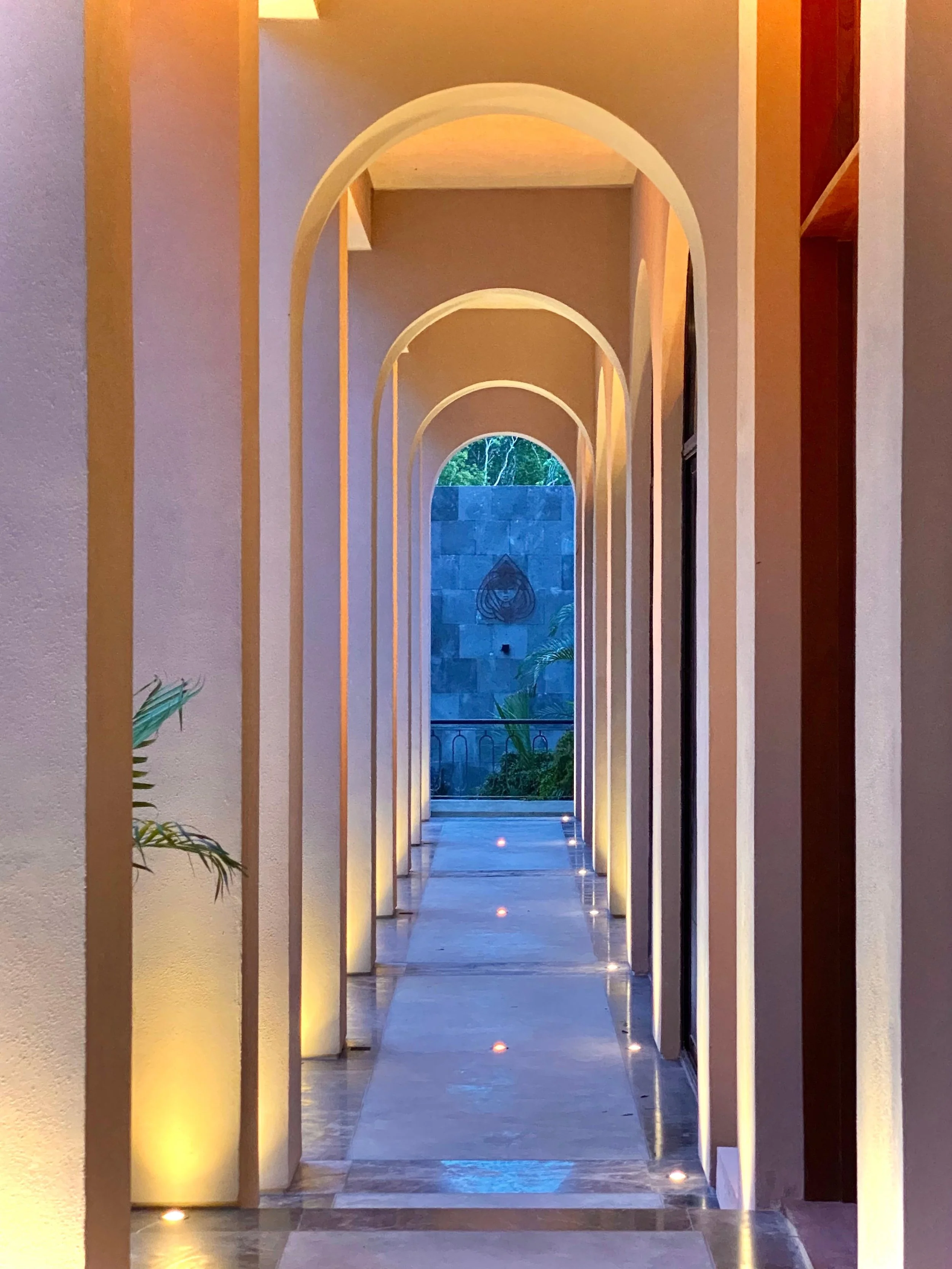 A hallway with multiple arches, illuminated by warm lights on the walls and floor, leading to a window with a view of greenery outside.