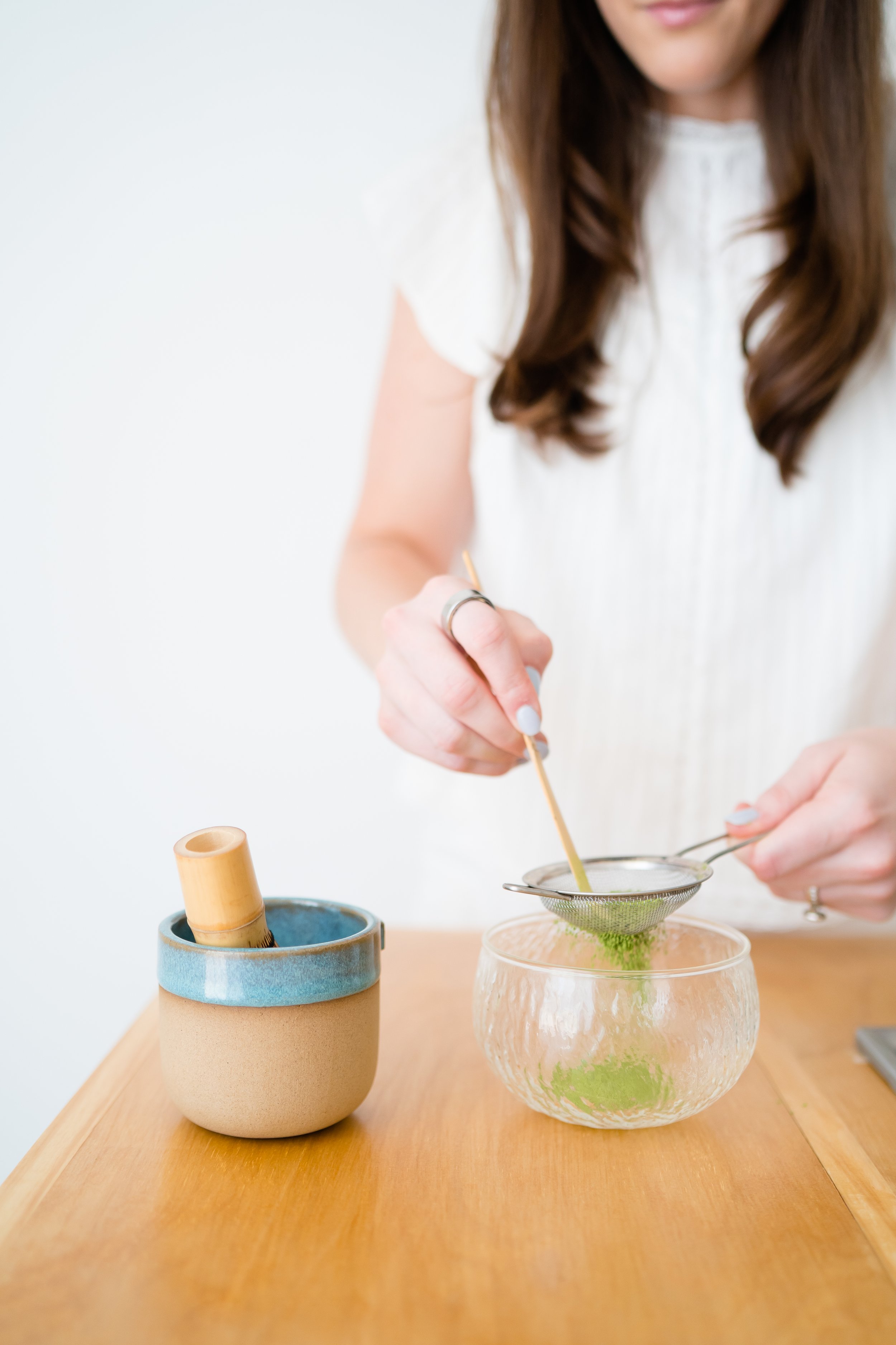 Close-up of a woman sifting matcha powder through a metal sieve into a glass bowl, with tea utensils on a wooden table