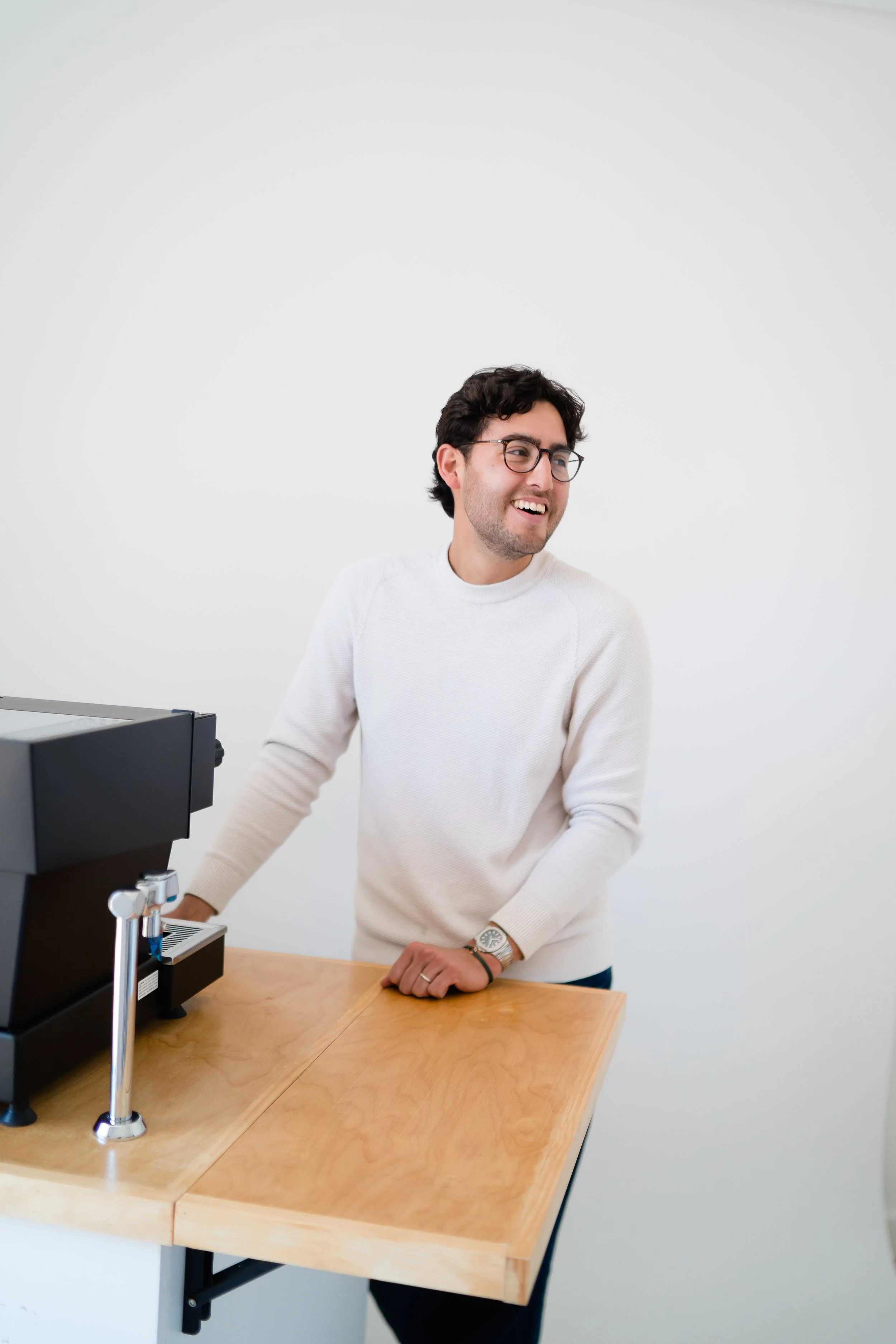 A man with dark curly hair, glasses, and a beard smiling while standing next to a coffee brewing machine on a wooden table in a minimal white room.