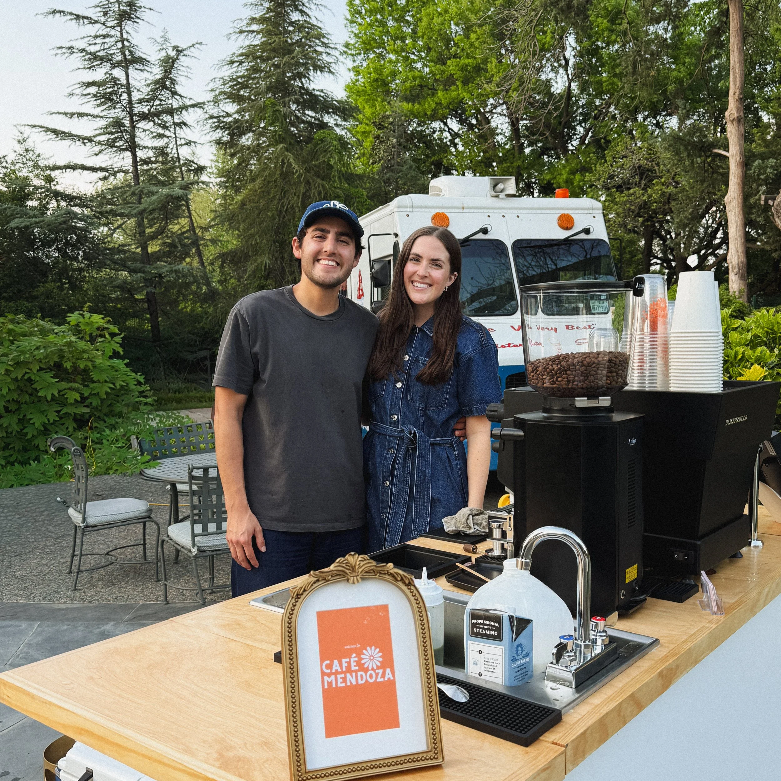 Two smiling people standing behind a coffee cart labeled Café Mendoza, with coffee supplies and equipment on the wooden counter, outdoors surrounded by trees.