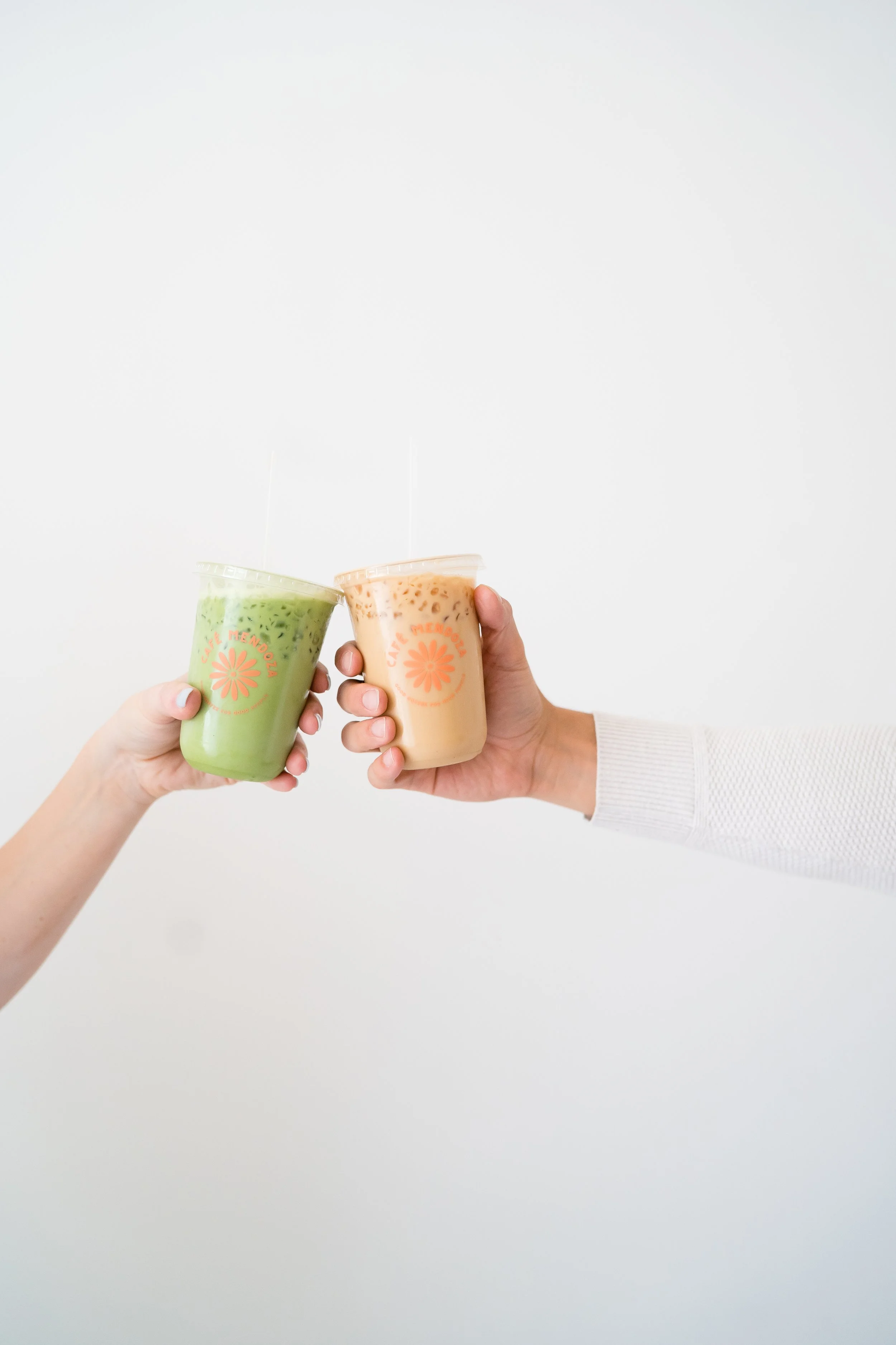 Two hands holding bubble tea drinks, one green and one peach, against a plain white background.
