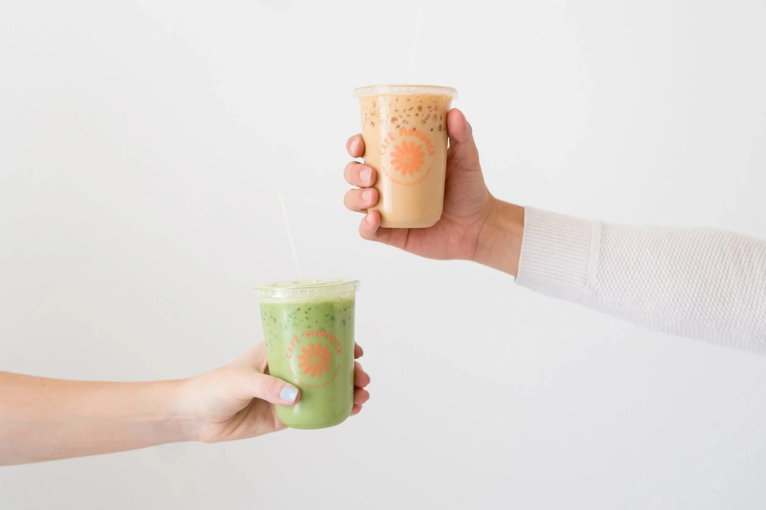 Three people are holding different beverages in transparent cups with orange logos. One person holds a green drink, while another holds a beige drink with a straw, against a plain white background.