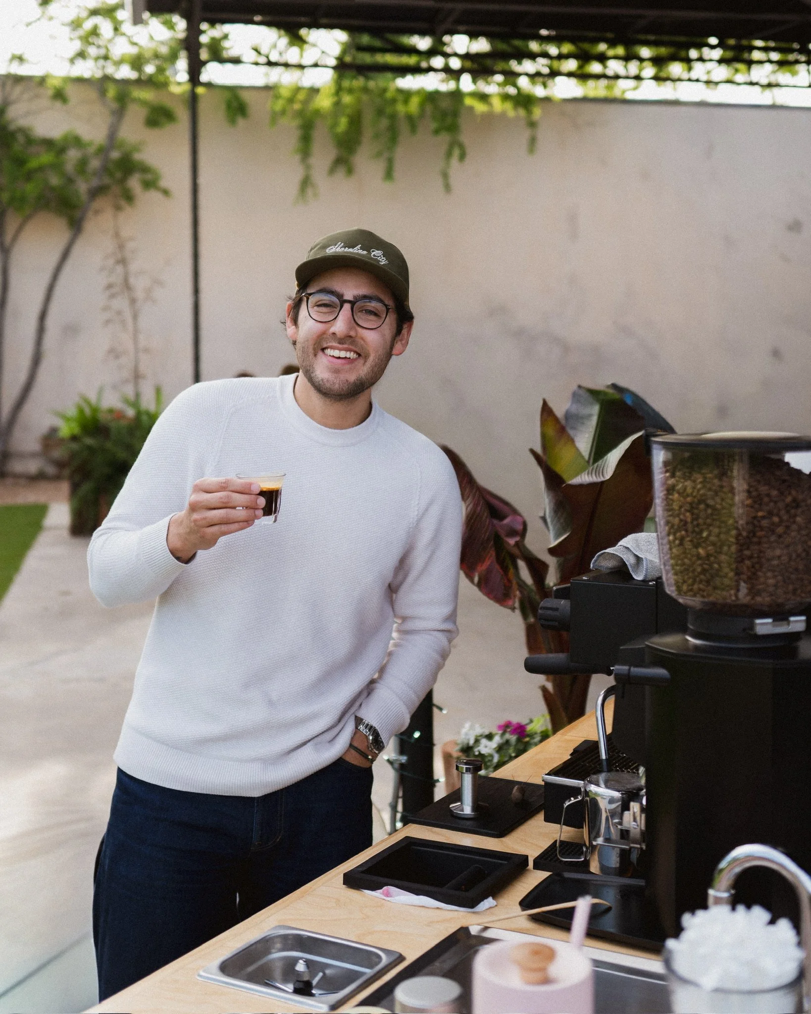 A man wearing glasses, a green baseball cap that says 'Shoreline City, and a white sweater, smiling while holding a glass of espresso. He is standing at a coffee cart with coffee brewing equipment, in an indoor space with plants and a beige wall.