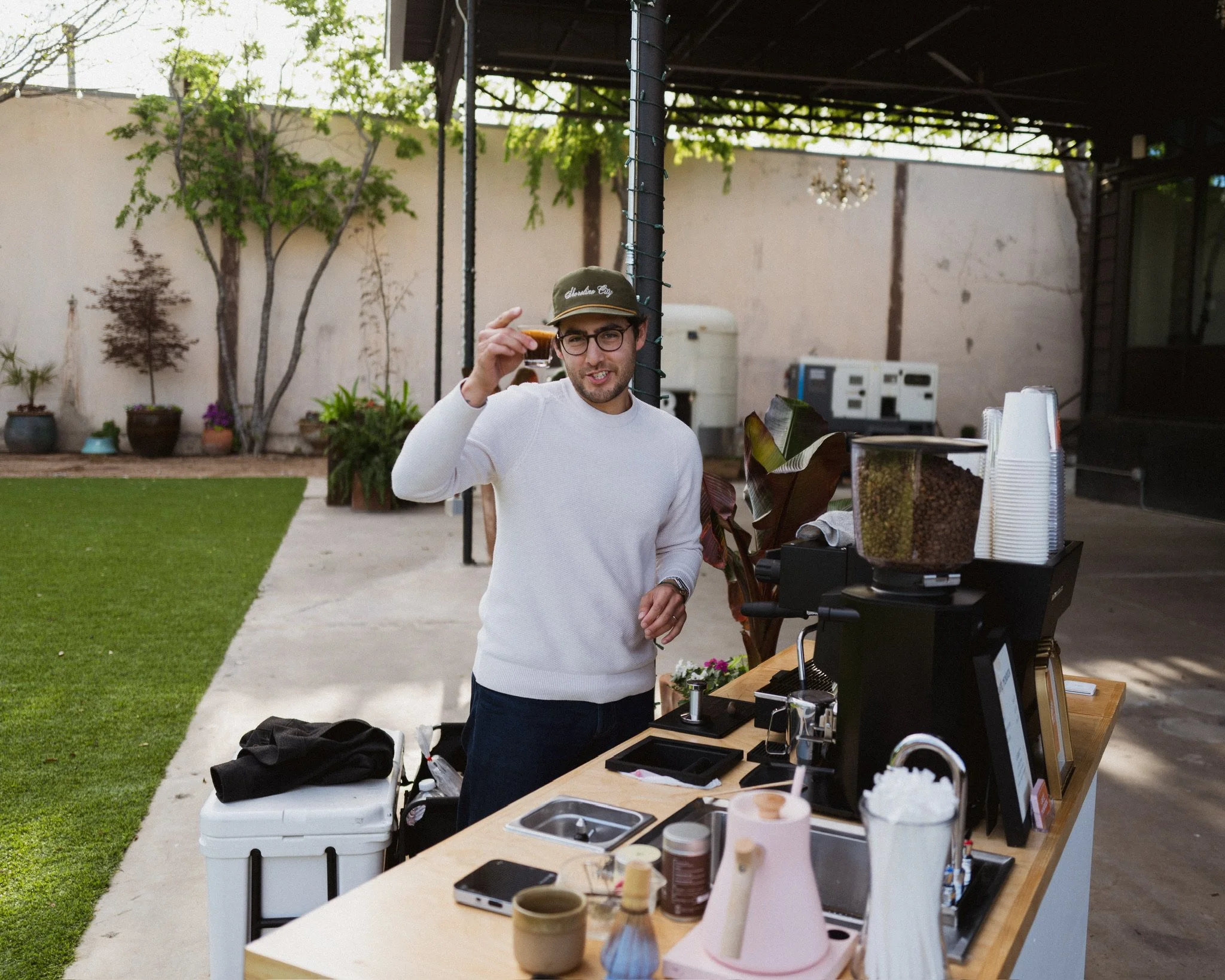 A man in a white sweater and glasses holding a small cup, standing at a table with various coffee supplies under a patio cover. In the background, there's a beige wall with trees and potted plants.