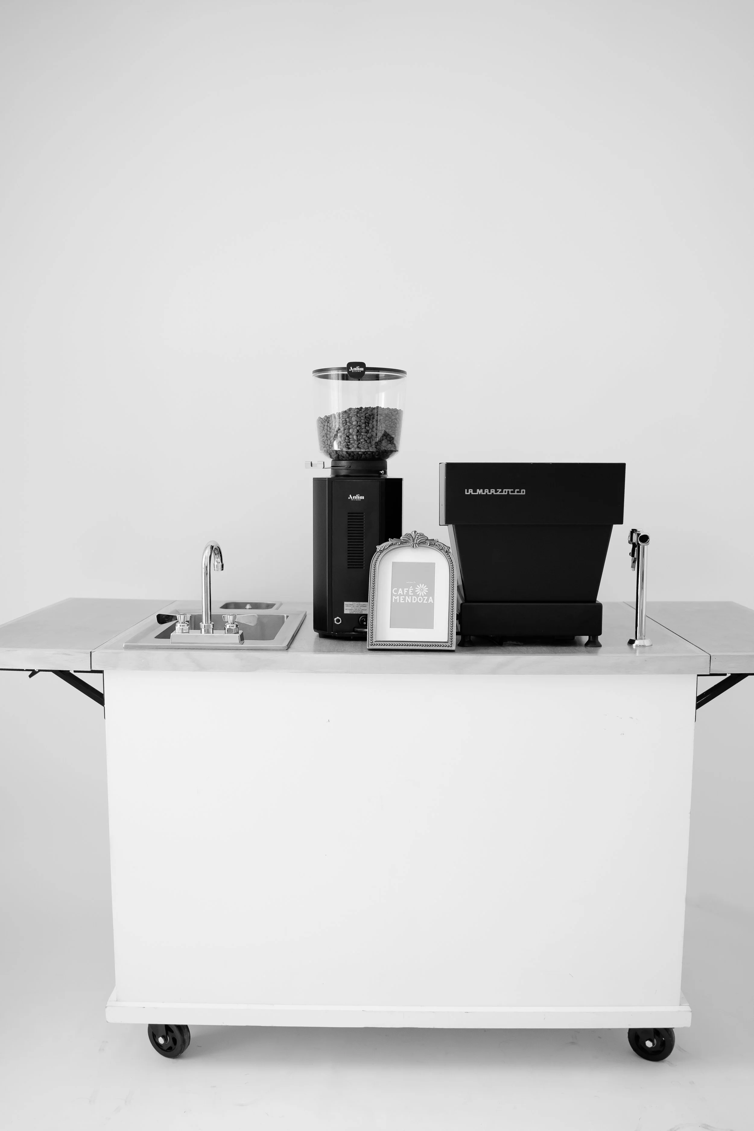 A white mobile counter with a built-in sink, a coffee grinder, a cash register, and a framed sign that reads 'Cafe Mendoza,' all against a plain white background.