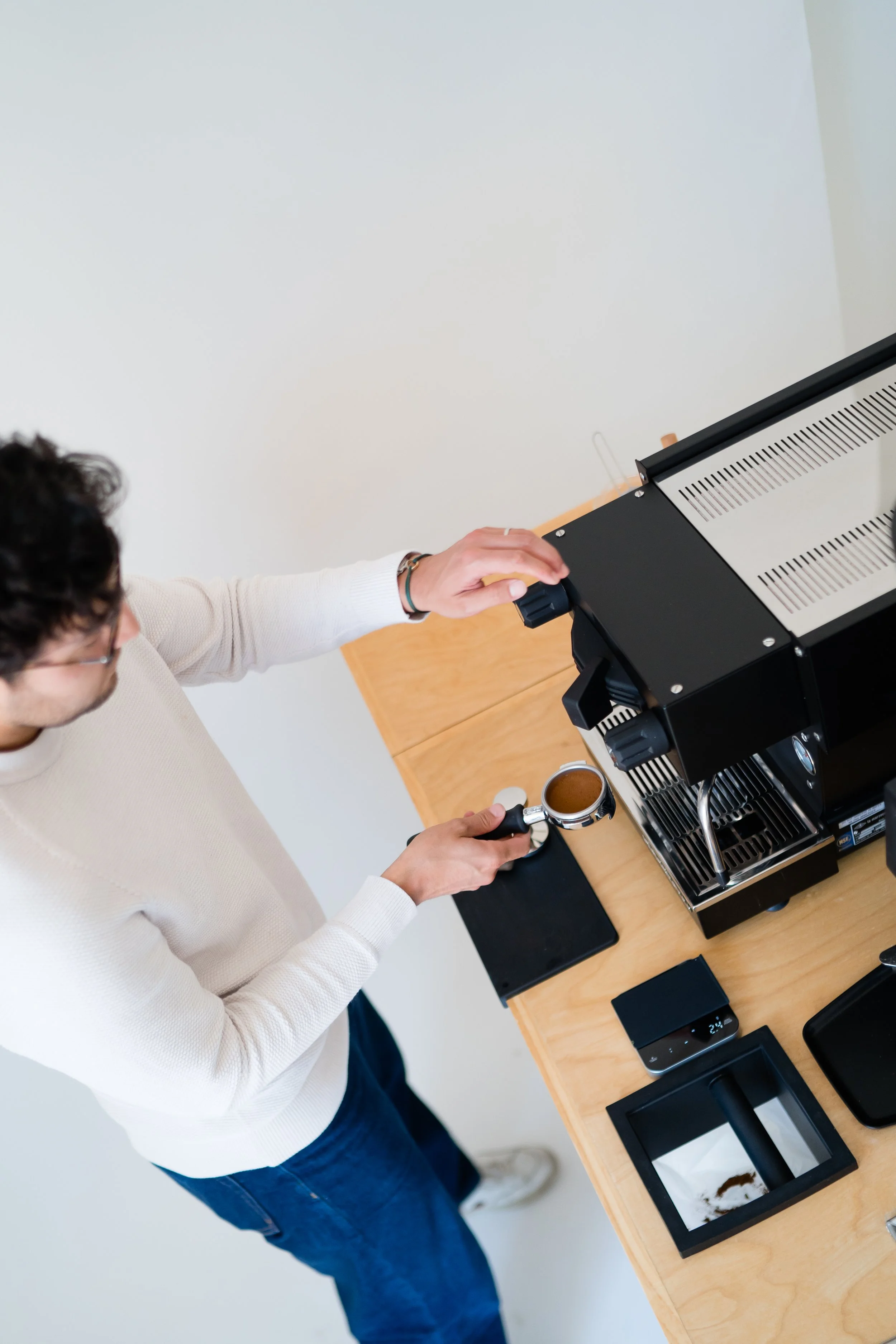 A man wearing a white sweater and glasses making coffee with a commercial espresso machine on a wooden table, with phones and other items nearby.