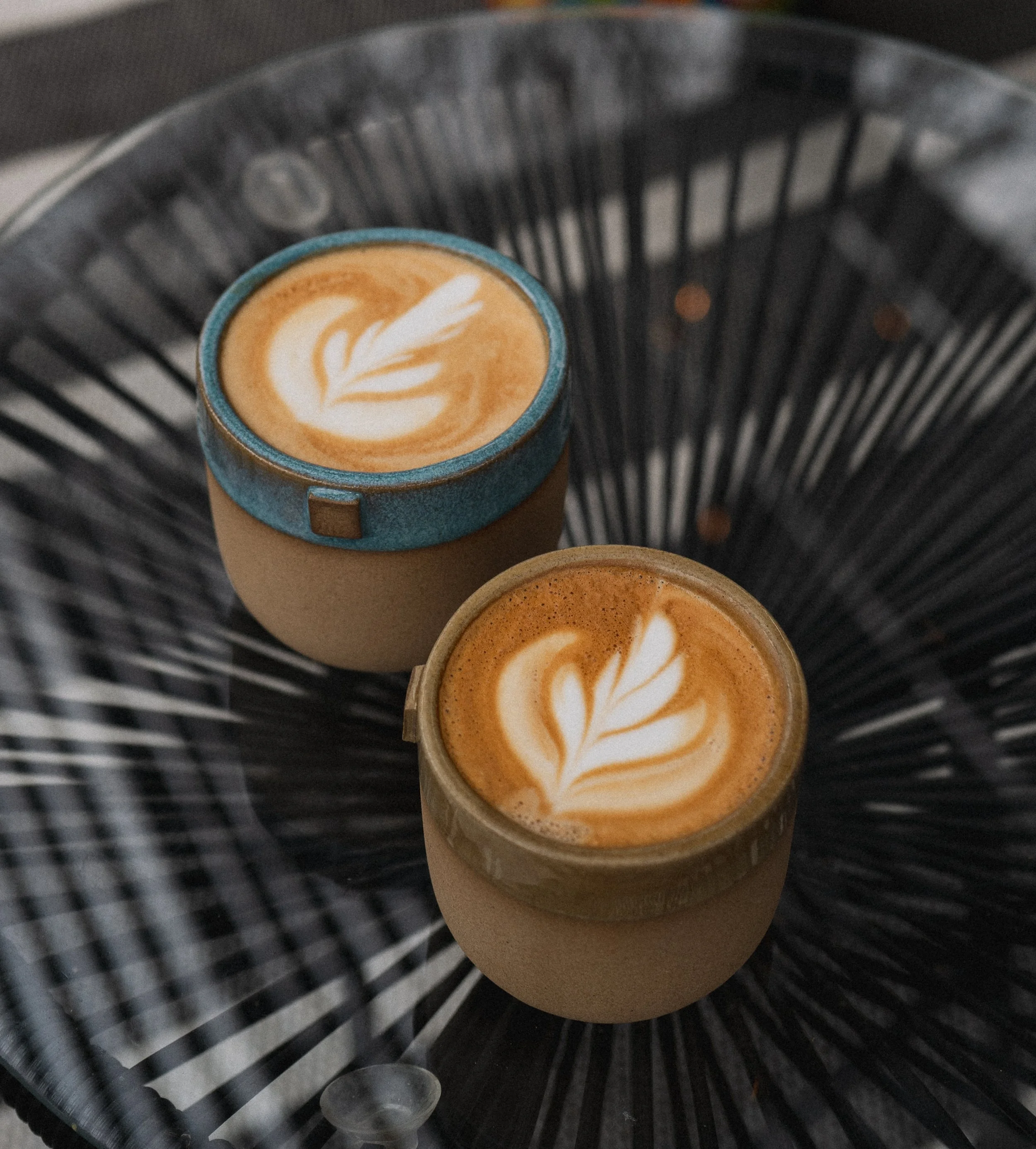 Two cups of coffee with latte art, placed on a black wire table.