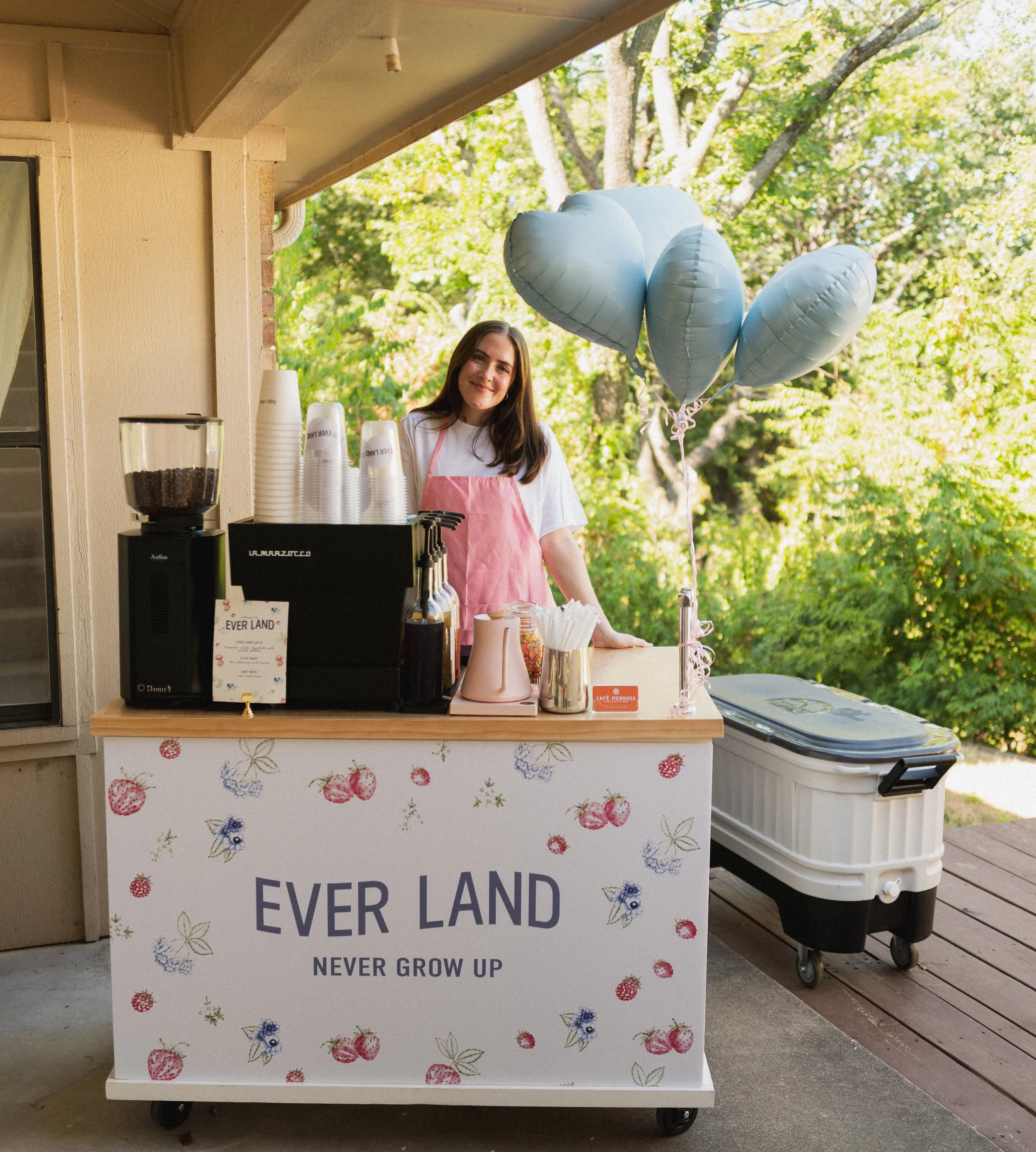 A person with long dark hair, wearing a pink apron, is standing behind a counter with balloons and various items, outdoors near trees. The counter has a sign with strawberries and flowers, reading "EVER LAND NEVER GROW UP".