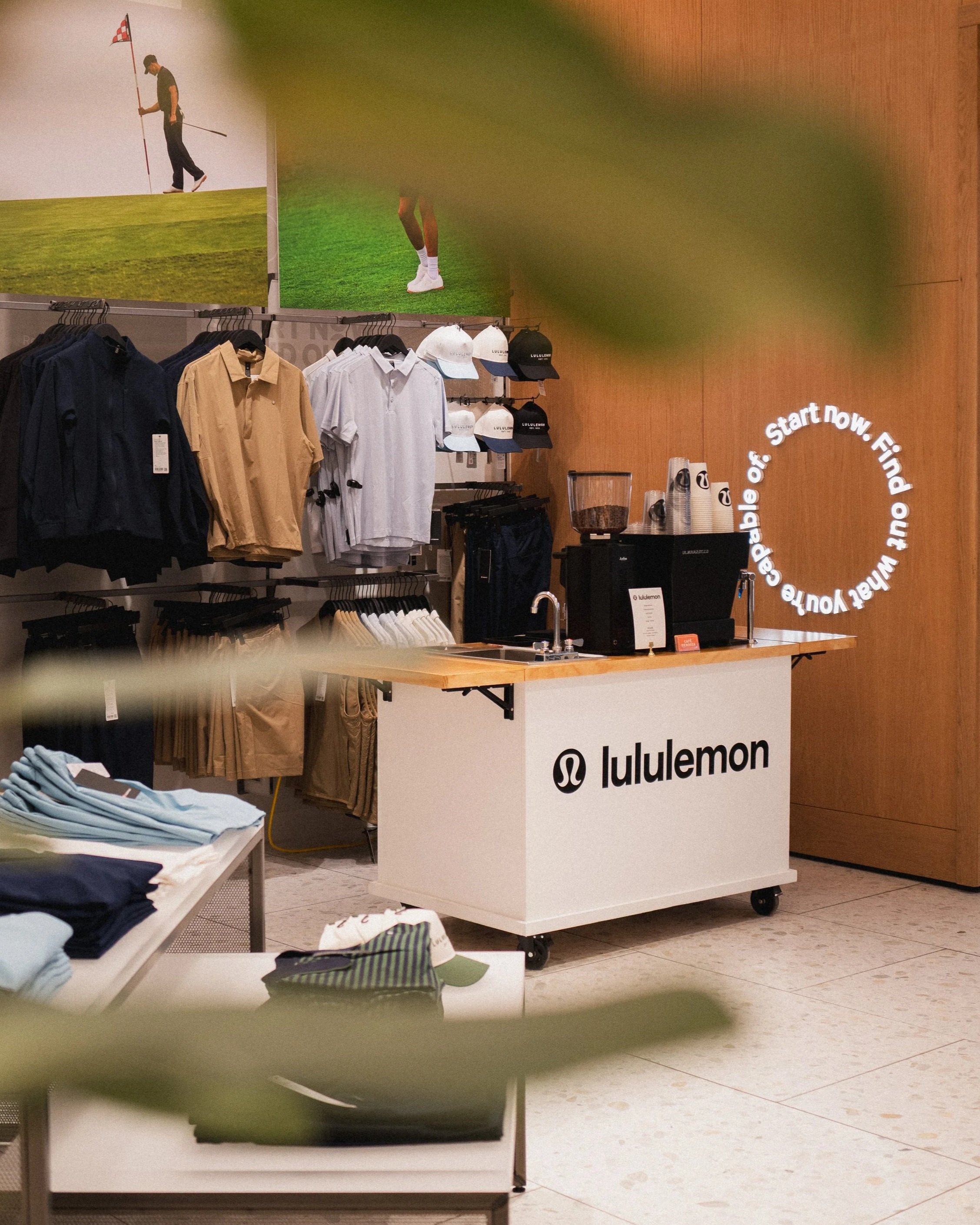 A small mobile coffee cart with a “lululemon” logo set up inside a retail clothing store, surrounded by neatly displayed apparel and hats, with a coffee machine and supplies on the counter for a brand activation event.