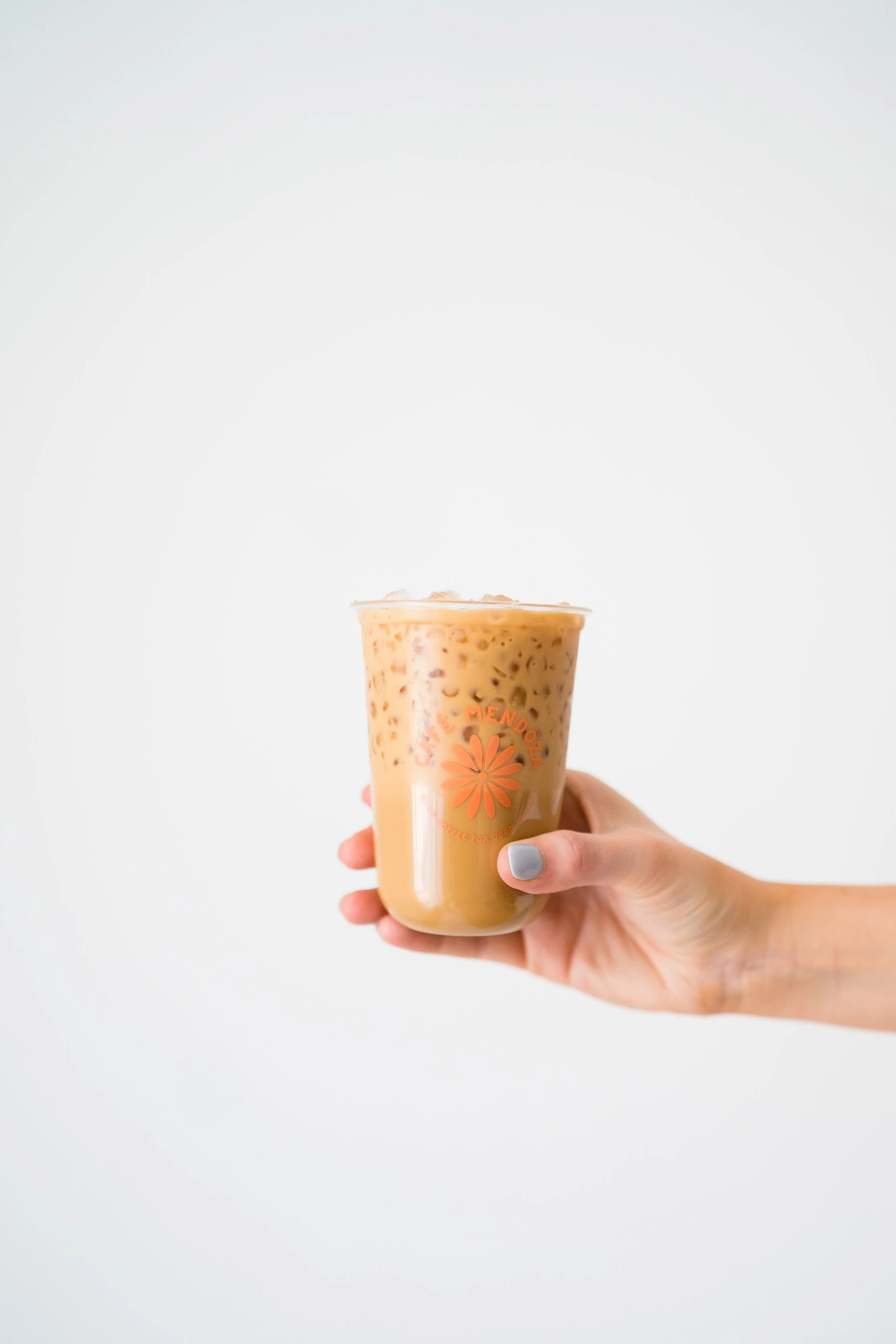 Hand holding a plastic cup of iced coffee or iced tea with a light brown color and ice cubes, against a plain white background.