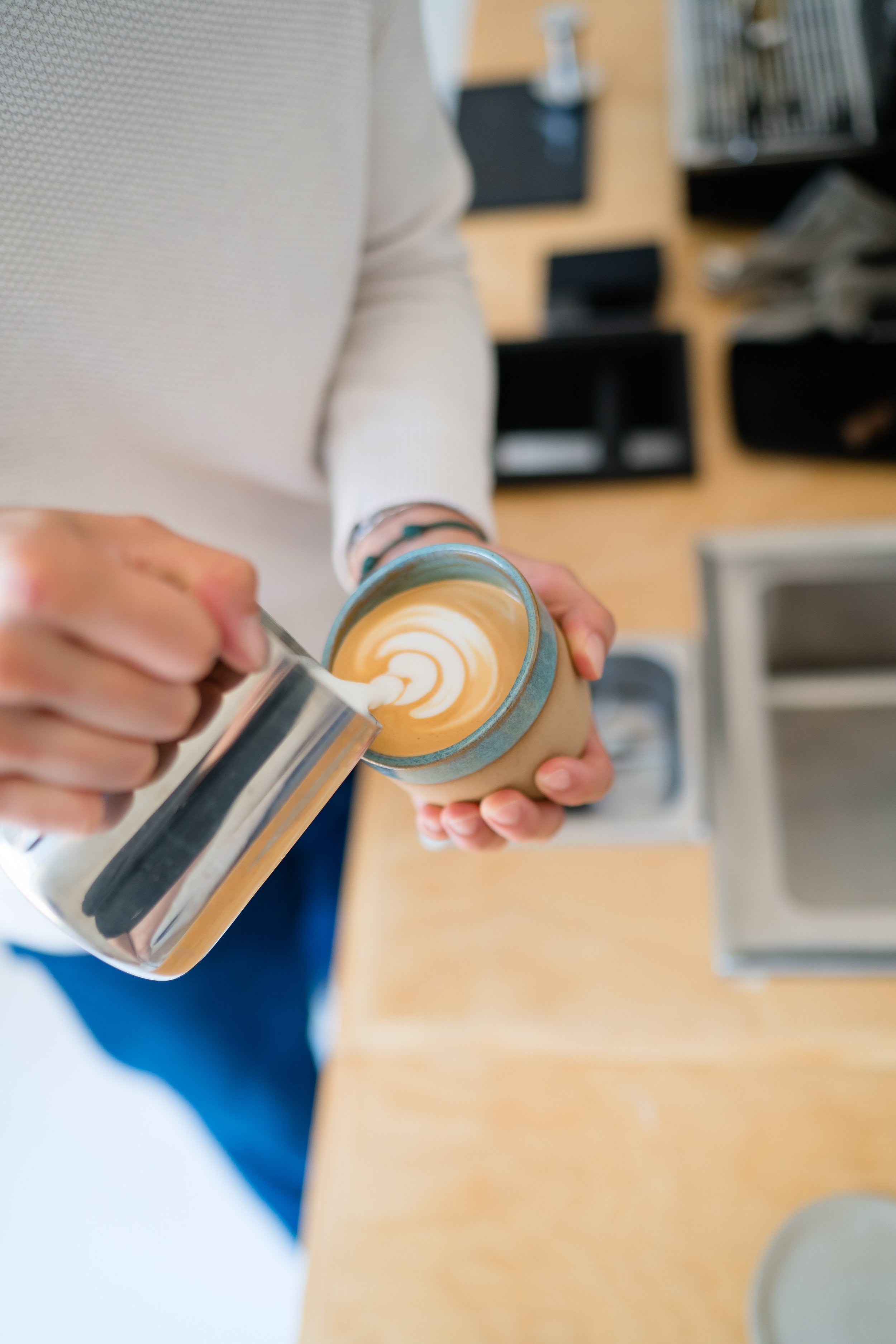 Person in a white sweater pouring steamed milk into a mug of coffee to create latte art.