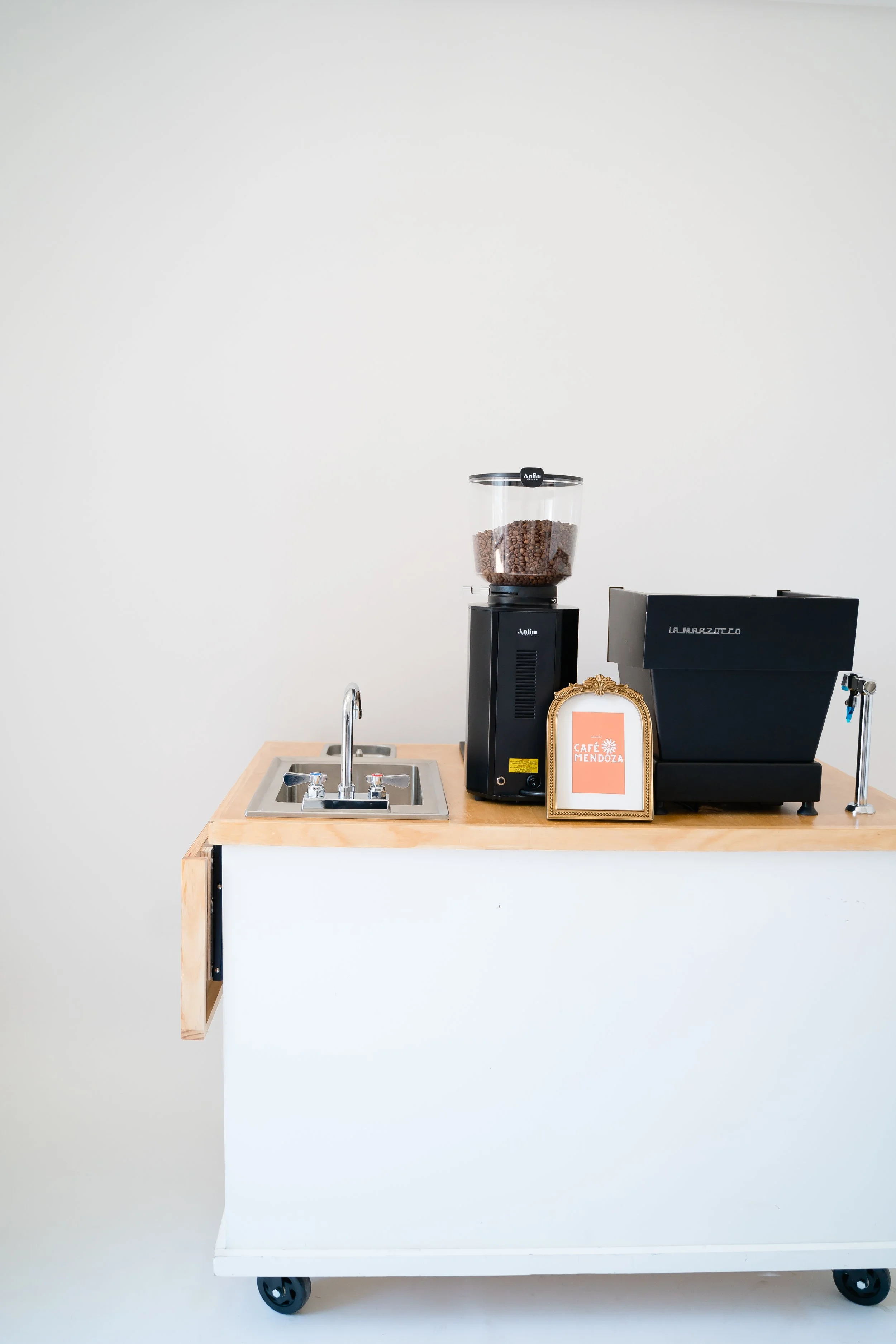 A coffee station with a small sink, a coffee grinder filled with coffee beans, and an espresso machine on a wooden counter with a white front, set against a plain white wall.