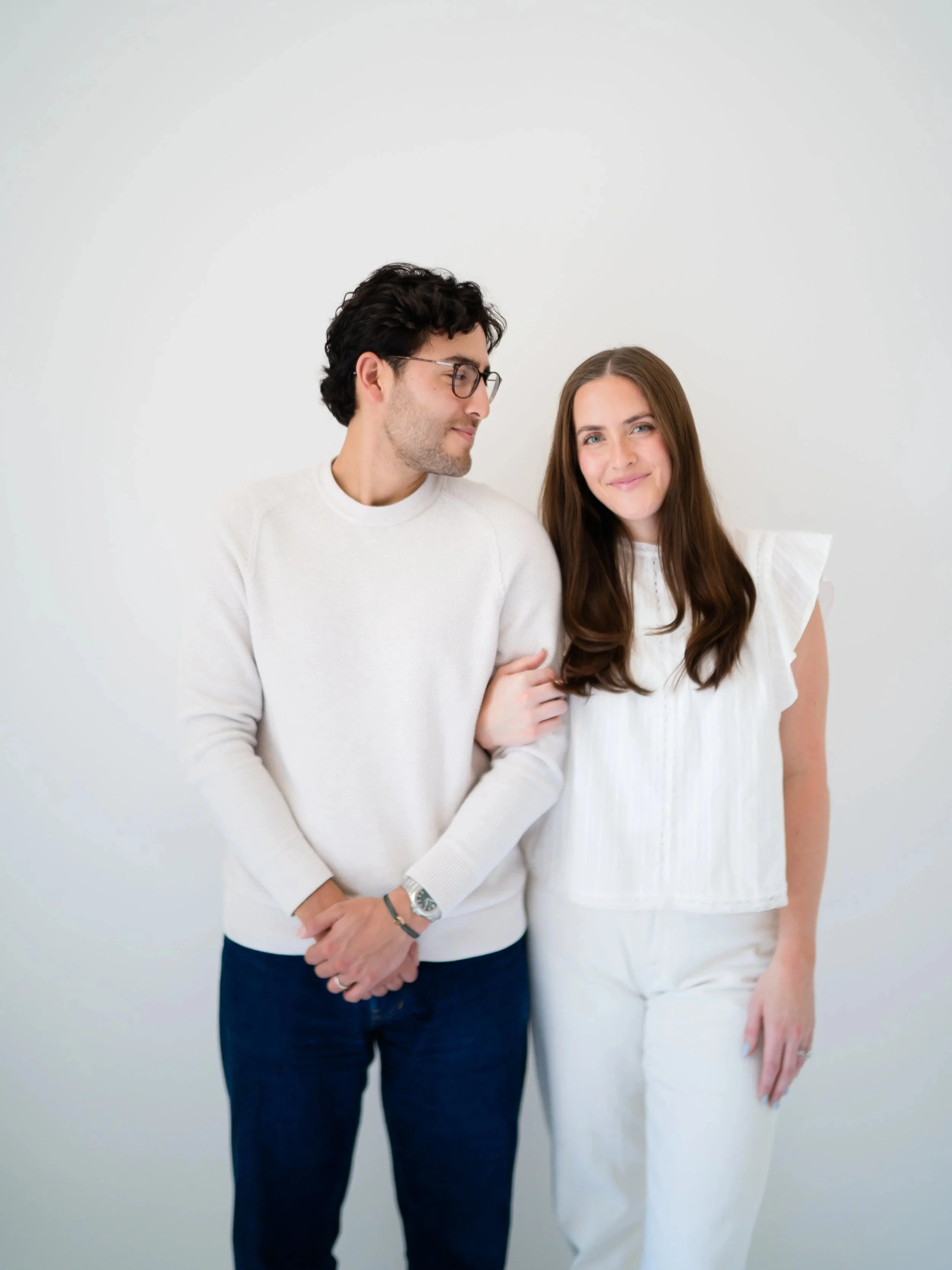 A man with dark hair, glasses, and a white sweater gently holding a woman's arm. The woman has long brown hair, a soft smile, and is wearing a white dress. They are standing against a plain white background, close to each other.
