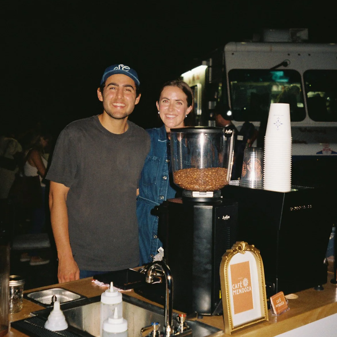 Two smiling people standing behind a coffee cart at night, with a coffee grinder and cups, and a sign that reads 'Café Mendoza'.