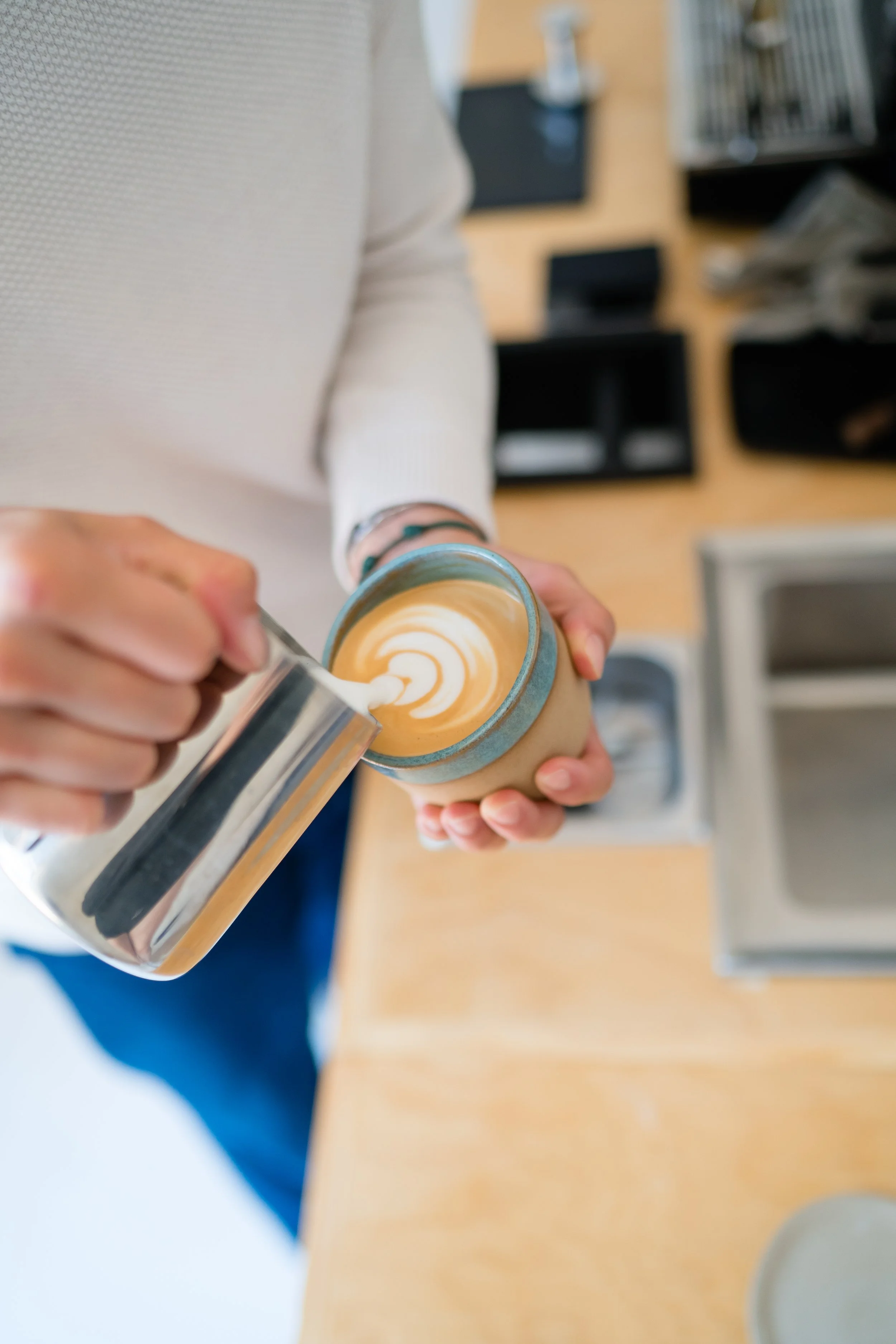 A person holding a mug of coffee while pouring frothy milk on top to make latte art, with a hand holding a container of milk and a latte cup on the counter.