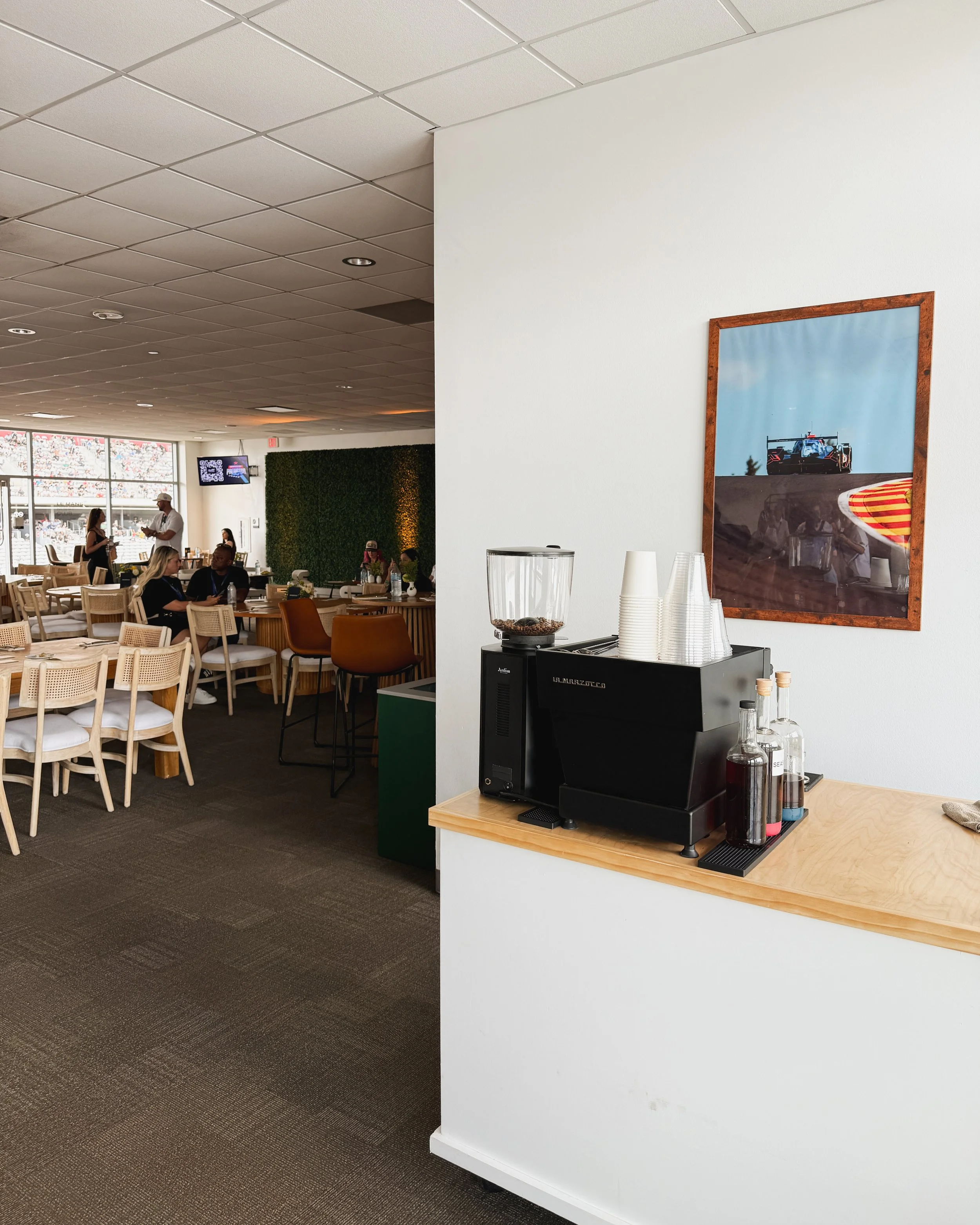A La Marzocco espresso machine and coffee grinder set up on a wood counter in a hospitality suite, with stacked cups and glass syrup bottles nearby, a framed photo of a race car on track hanging on the wall, and a lounge area with cane chairs.