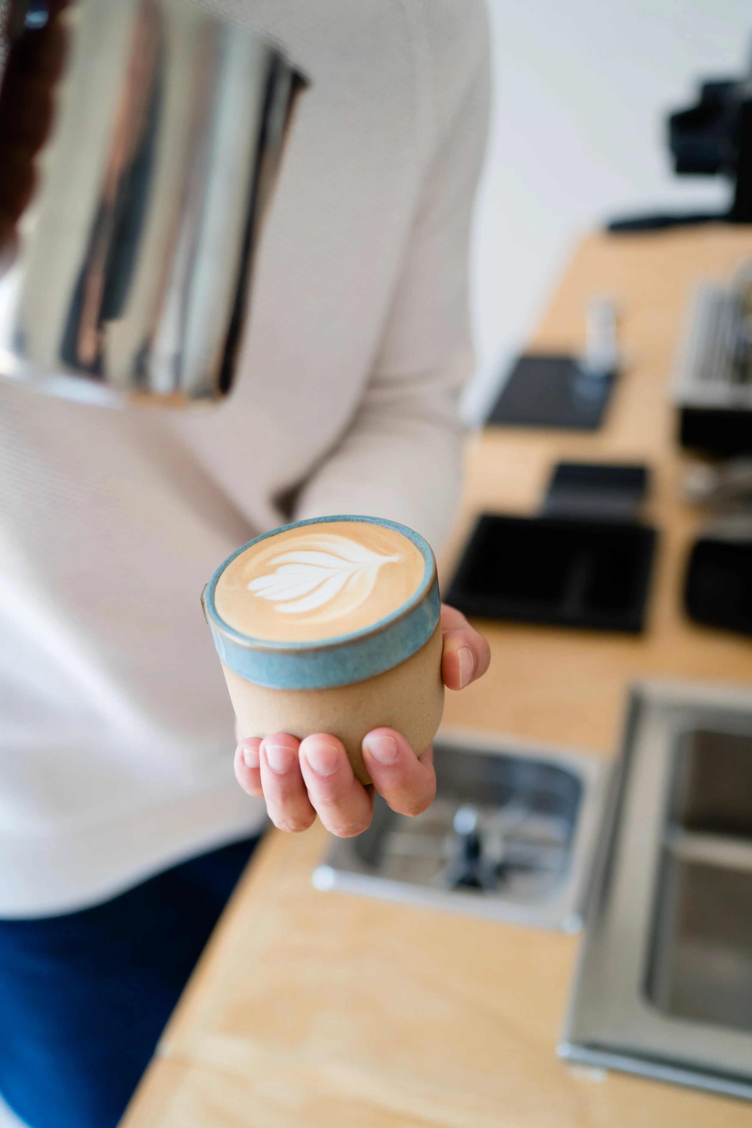 Person holding a cup of coffee with latte art in a café or coffee shop, with bookshelves and a coffee machine in the background.