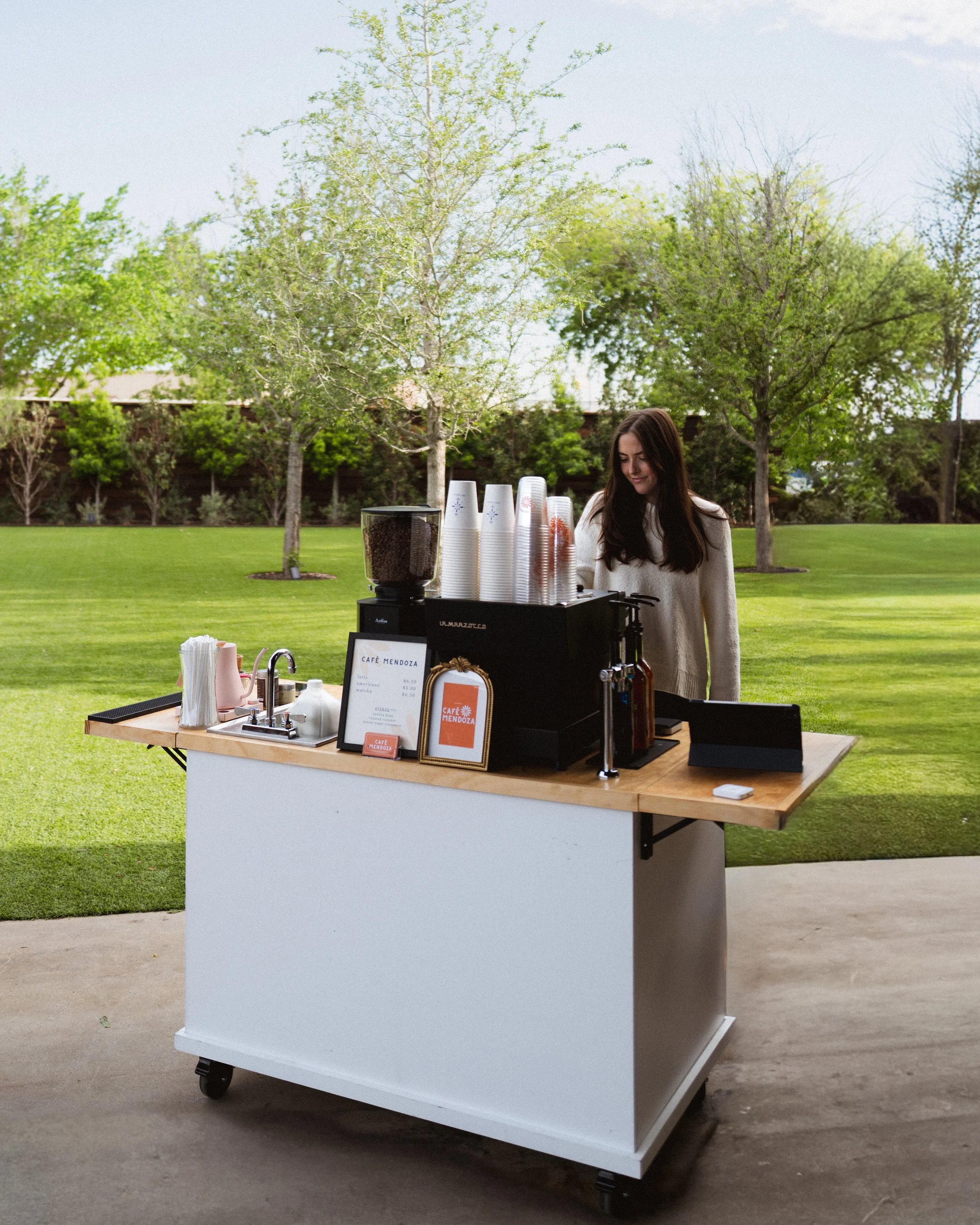 A woman in a white sweater serves coffee at an outdoor coffee cart with a wooden countertop, cups, and a sign, in a park with green trees and grass.