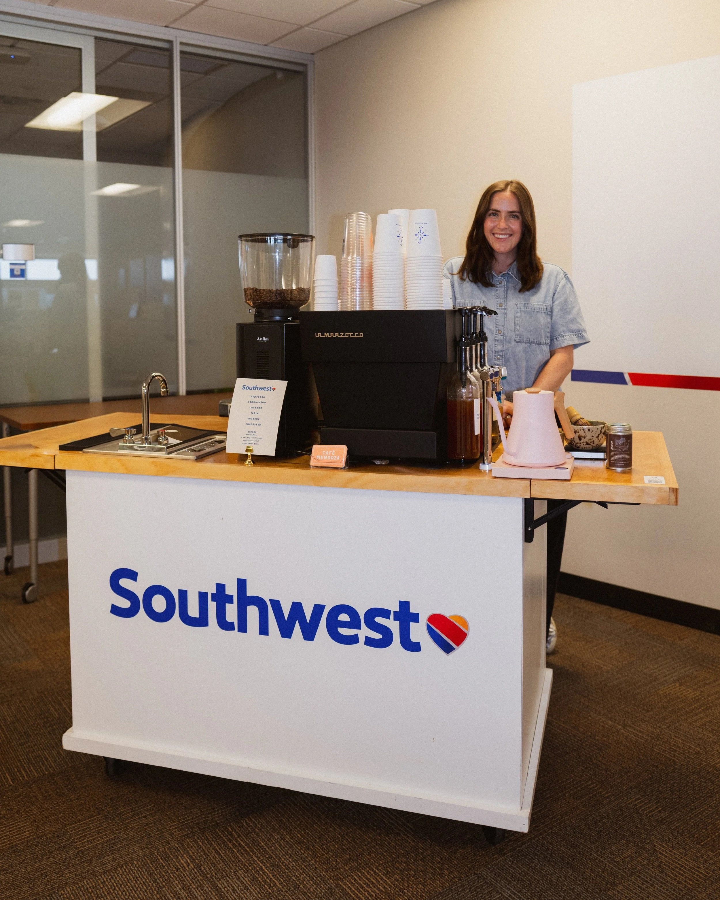 A mobile coffee cart branded with the Southwest logo set up indoors, with a barista standing behind an espresso machine, cups, and syrup bottles, serving coffee at a corporate event.
