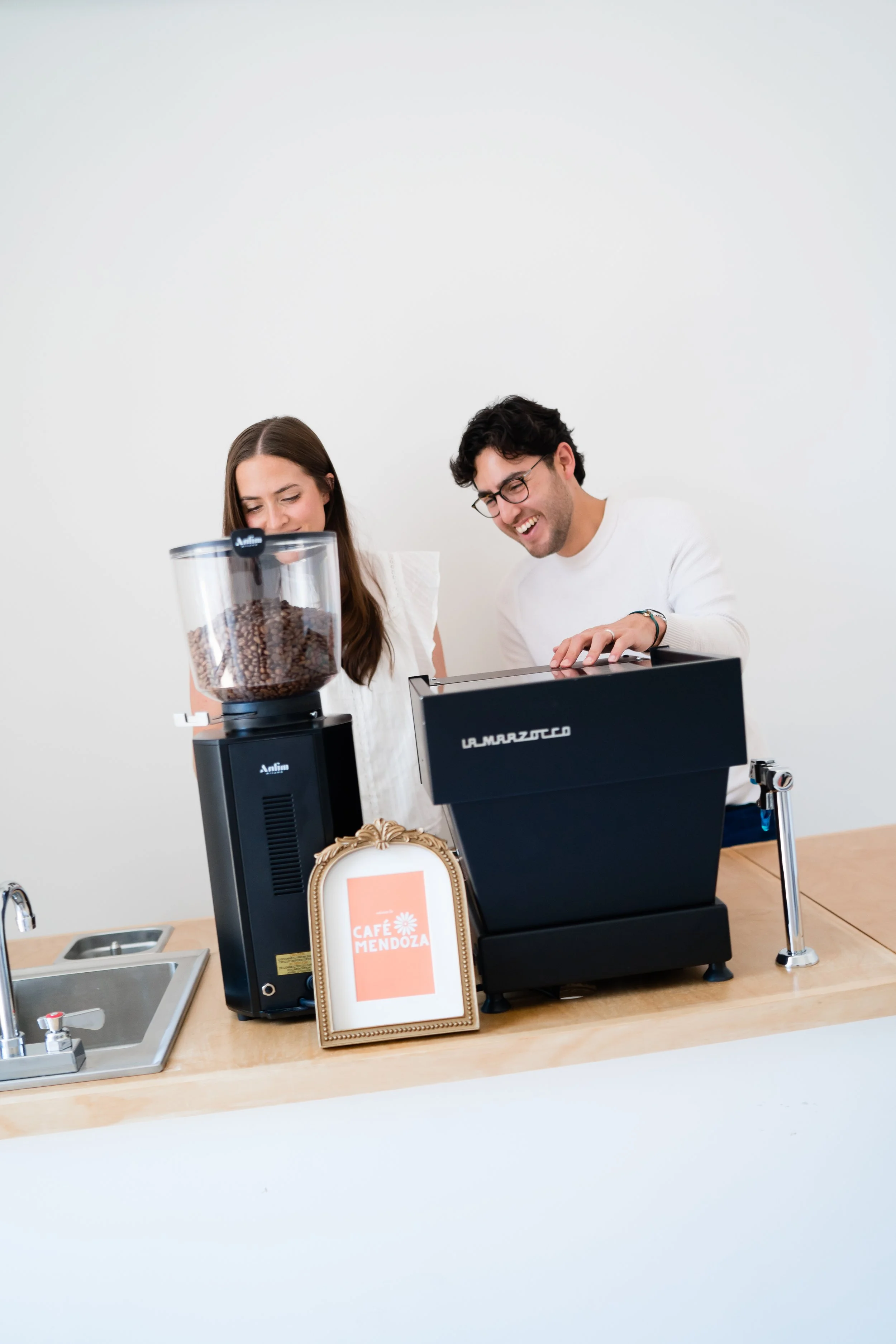 Two people making coffee behind a counter at Cafe Mendoza, with a coffee grinder, a framed menu, and a sink visible.
