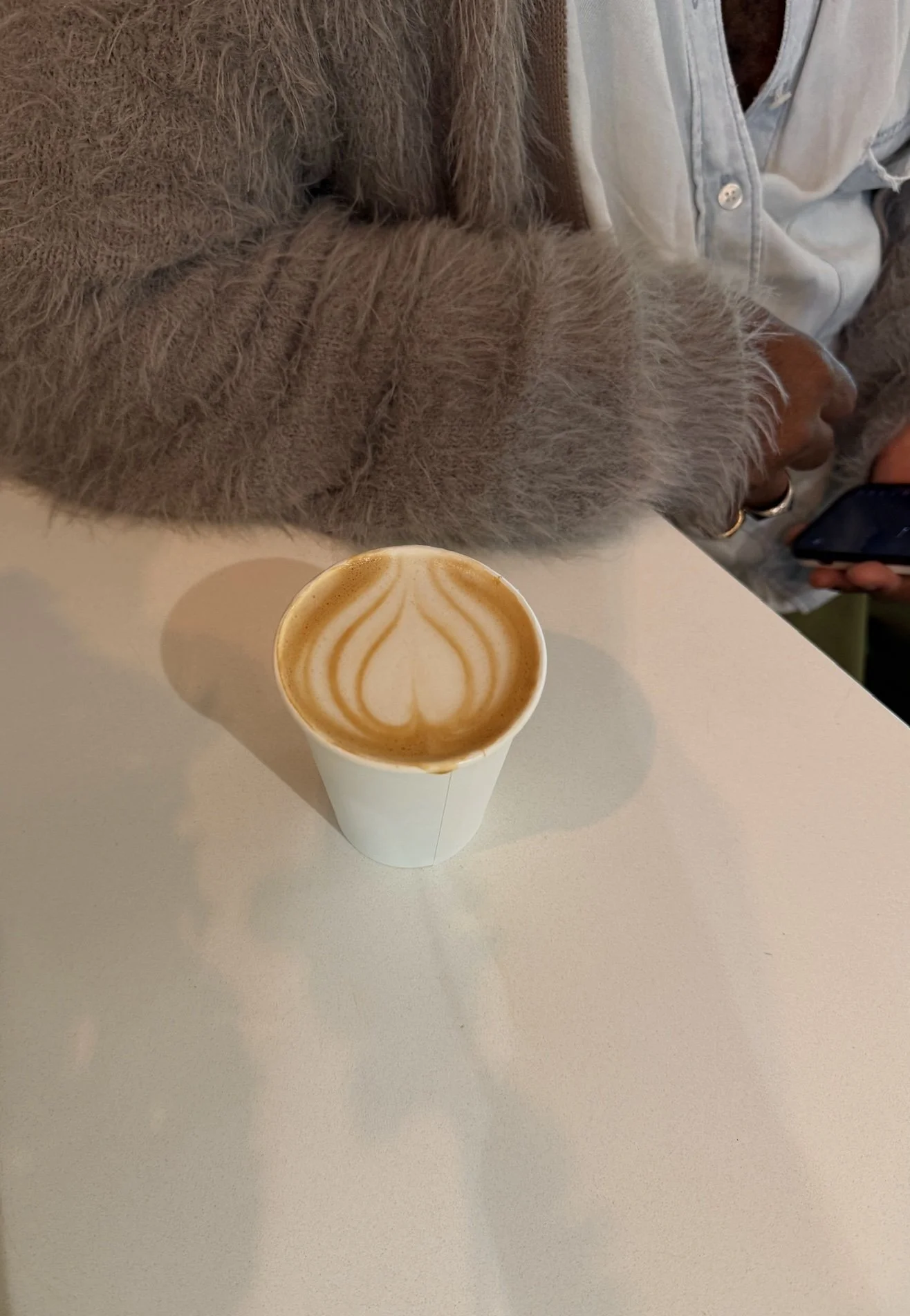 A cup of coffee with latte art on top placed on a white table next to a person's arm and torso, with part of their hand and phone visible.