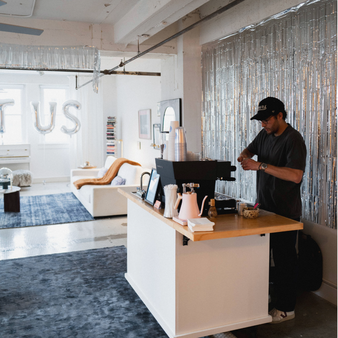 A person preparing coffee at a coffee cart stand inside a trendy, well-decorated lounge or studio space with cozy furniture, books, and shiny metallic decorations.