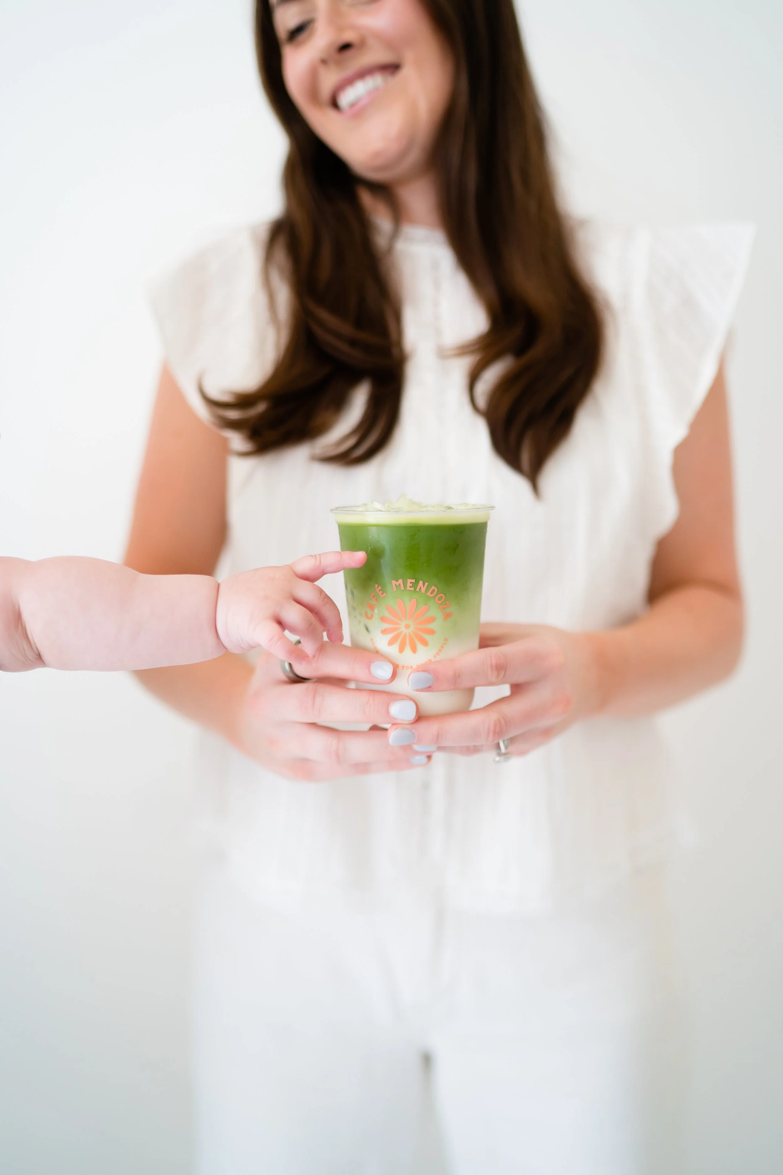 A woman with long brown hair smiling while holding a cup of green matcha latte with cream on top, and a child with small hand reaching out to touch the cup.