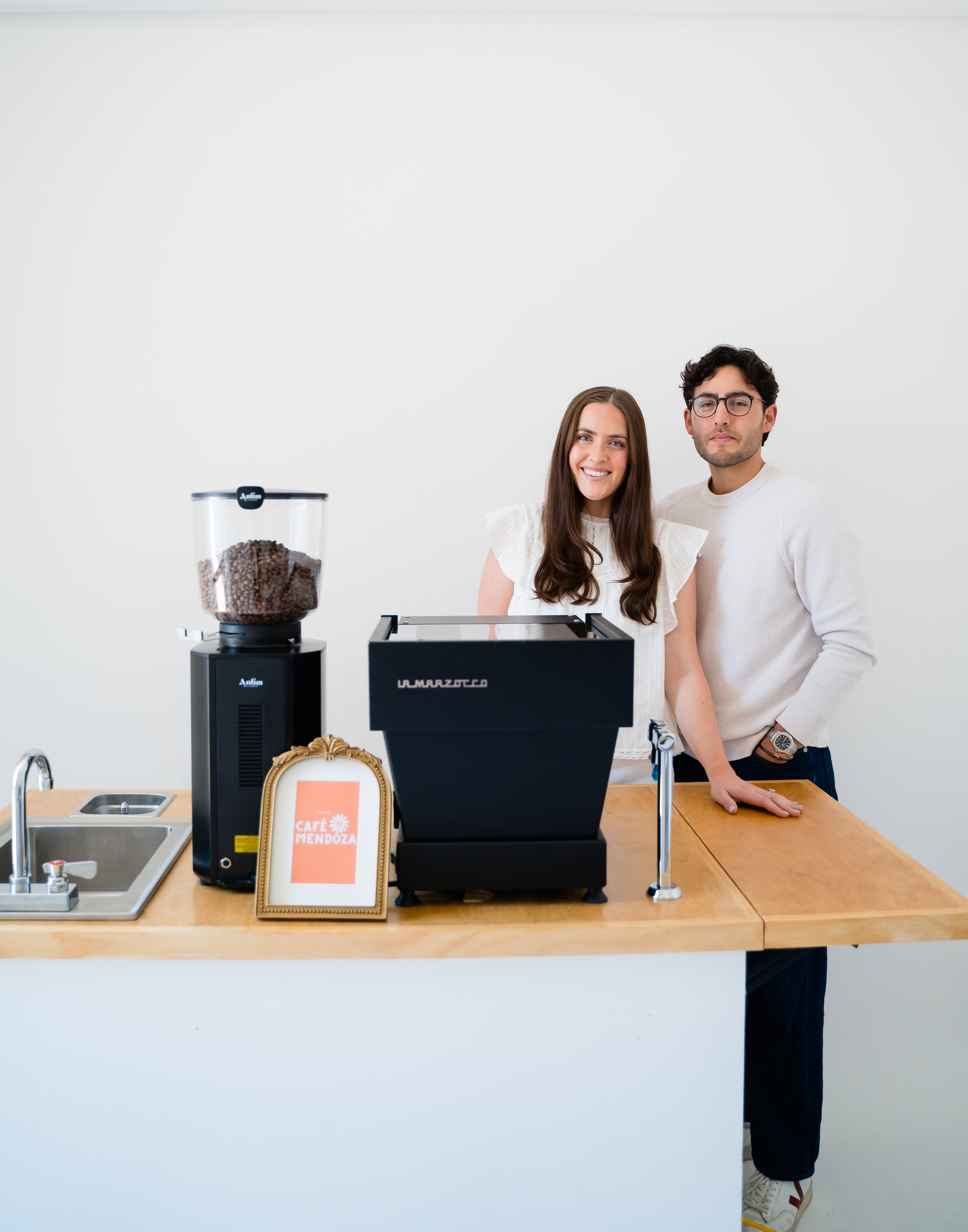 Young man and woman standing behind a coffee machine at a cafe, smiling, with a white wall in the background.
