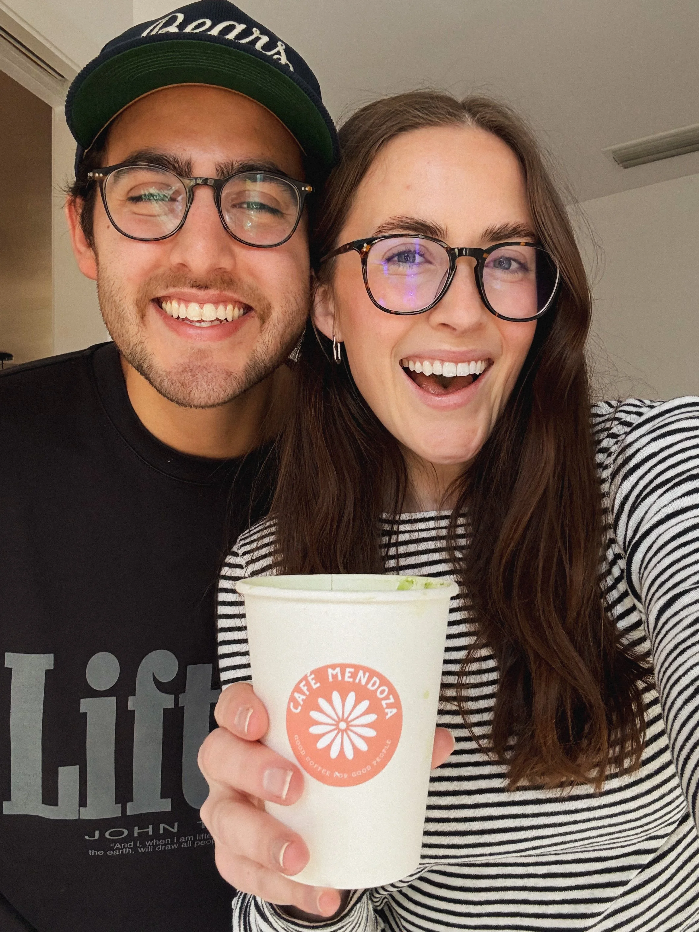 A smiling man and woman wearing glasses holding a coffee cup with a logo from Café Mendoza, inside a room with neutral walls.