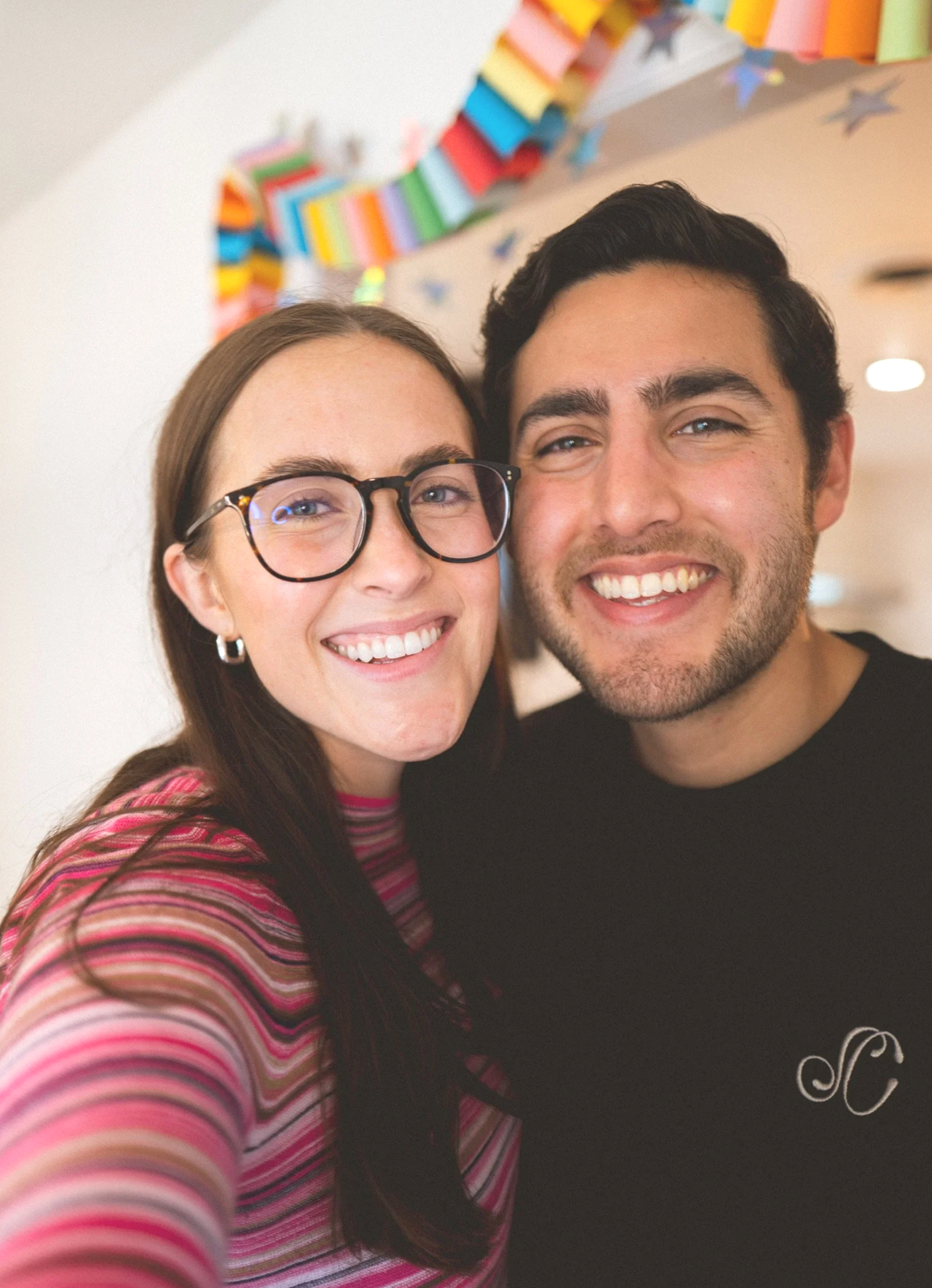 A smiling woman with glasses and long brown hair, wearing a pink striped shirt, taking a selfie with a smiling man with dark hair, wearing a black shirt. Behind them is a wall with colorful paper decorations and star-shaped ornaments.