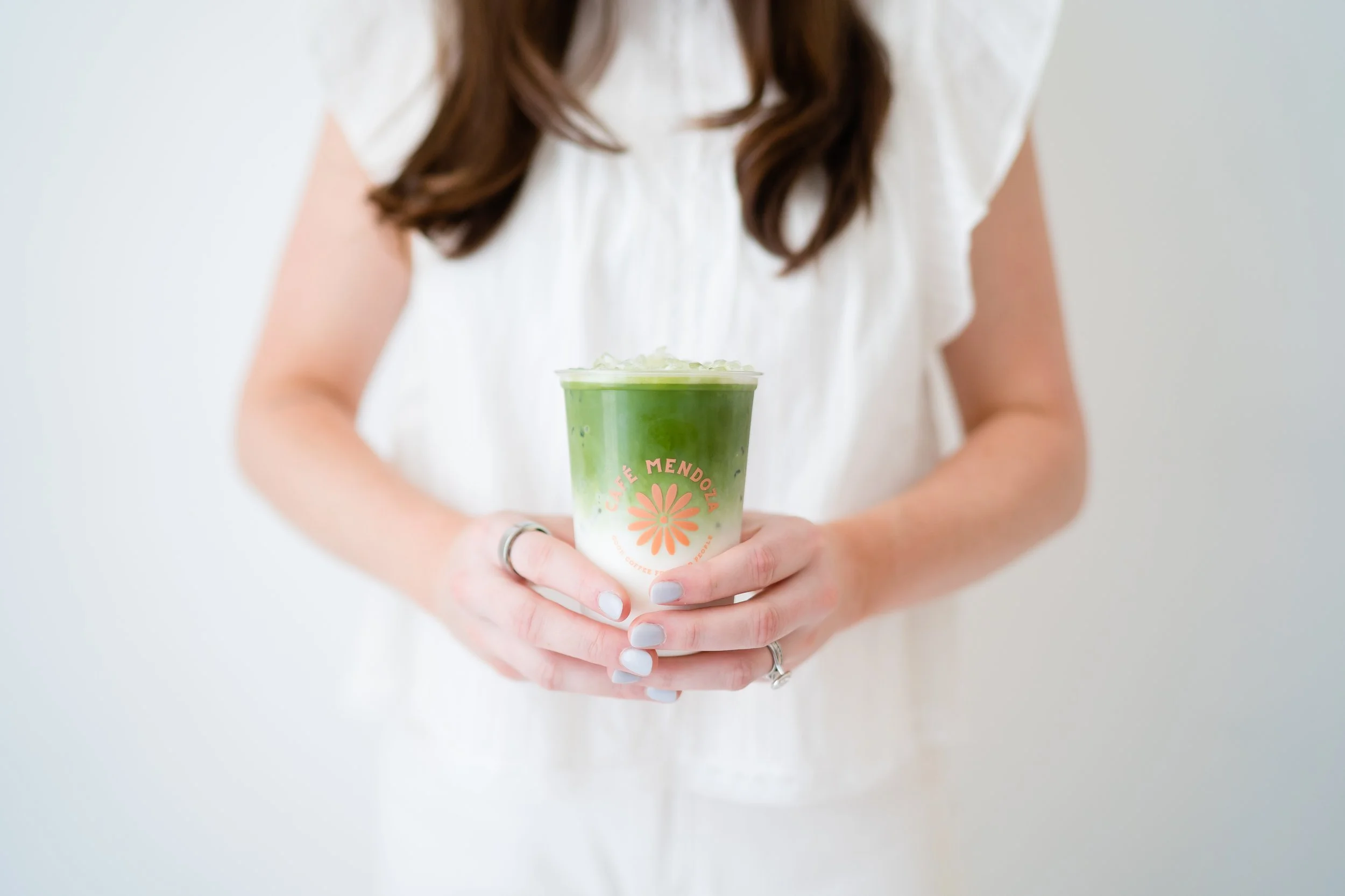 A woman with long brown hair, wearing a white dress, holds a green beverage in a clear cup with a floral logo and pink text that says "Café Mendoza."