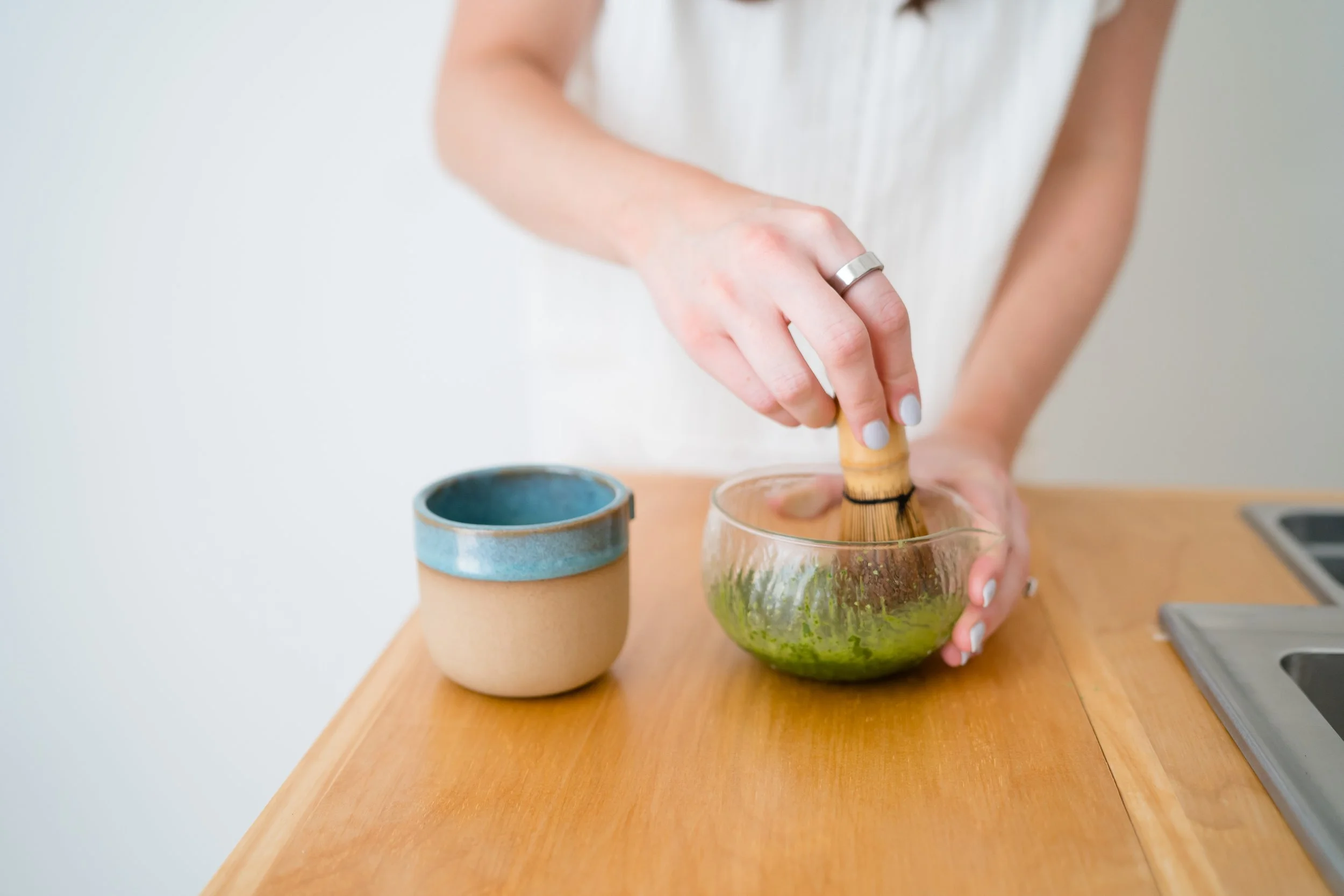 Person wearing a white top is using a brush to clean a glass bowl filled with green plants on a wooden table.