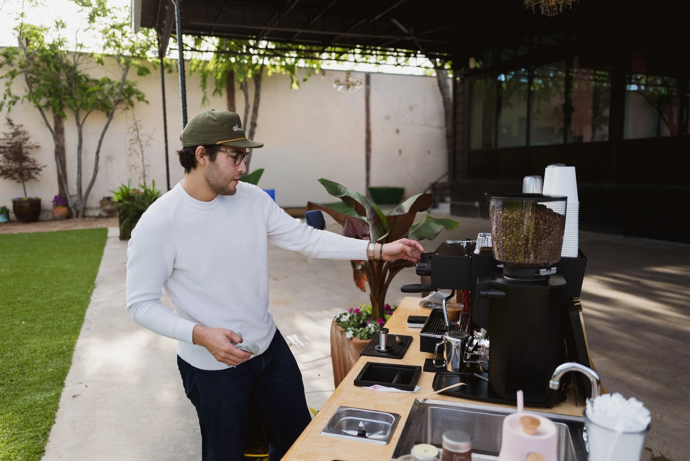 A man with glasses, wearing a white sweater and a green cap, making coffee at an outdoor coffee cart. There are plants, trees, and a patio in the background.