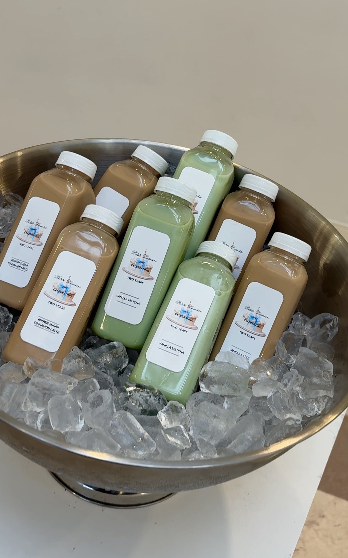 A silver bowl filled with ice contains bottled lattes with white caps, some labeled as vanilla matcha and others as cinnamon latte, arranged on a white surface.