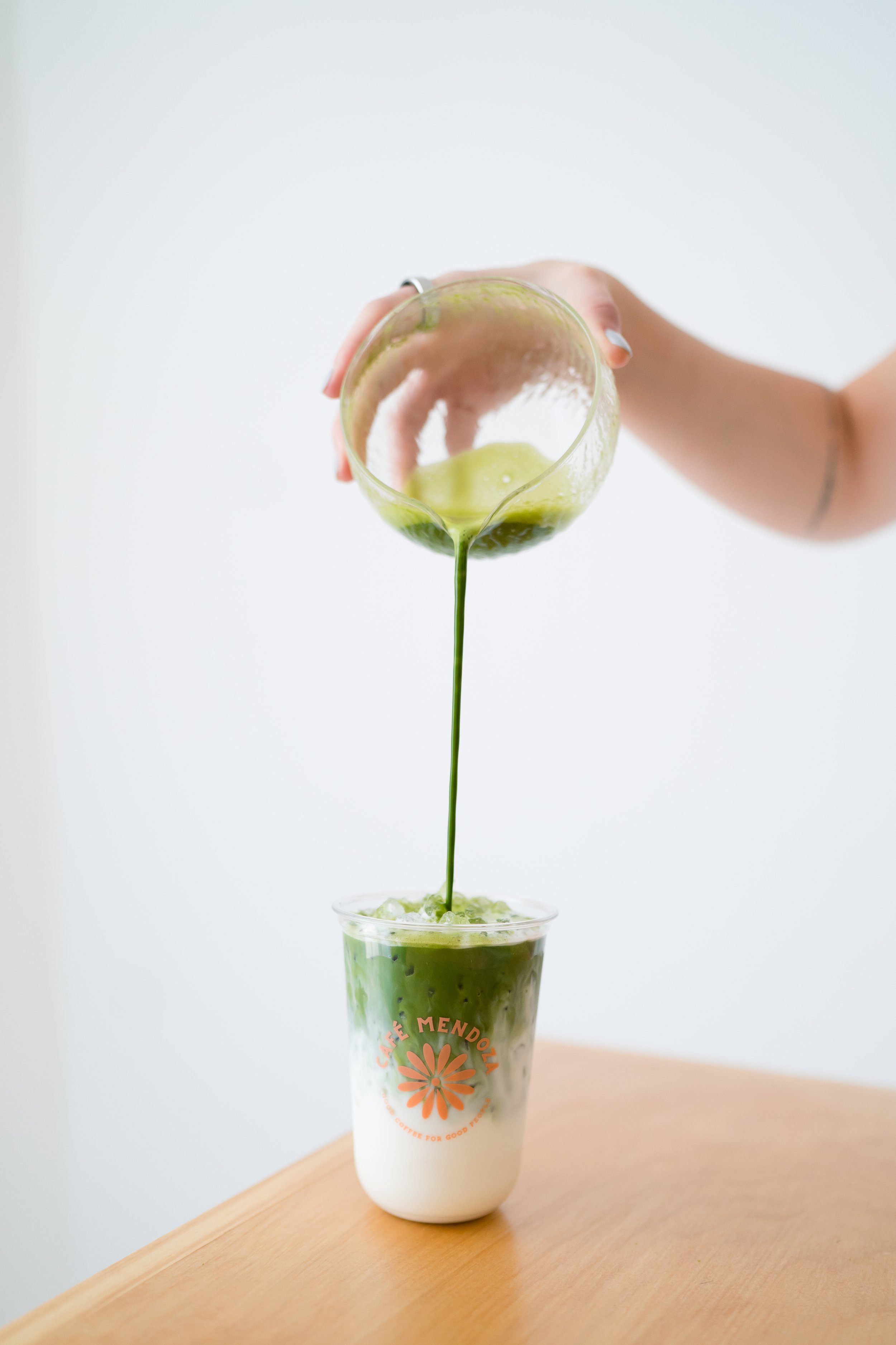 A hand pouring a green matcha latte from a glass pitcher into a clear cup with the Café Mendolín logo on it, containing layers of matcha and milk.