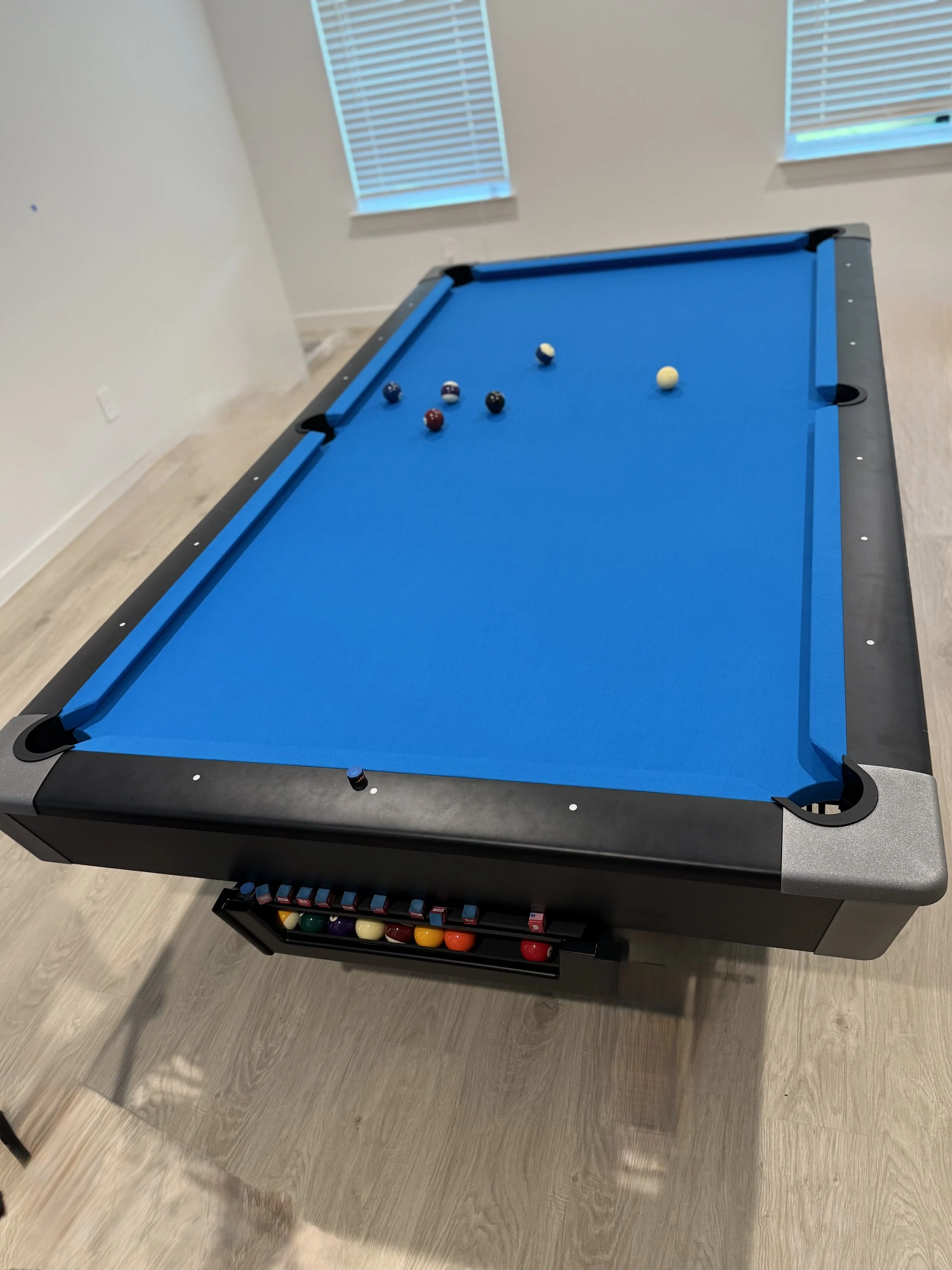 A blue-felt pool table with billiard balls scattered on it. The table has black edges and appears to be in a room with light wooden floors and white walls. Two windows with blinds are visible in the background.