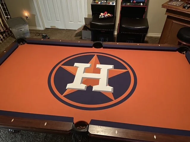 A pool table with the Houston Astros logo in the center, featuring a white 'H' inside a navy star on an orange background.