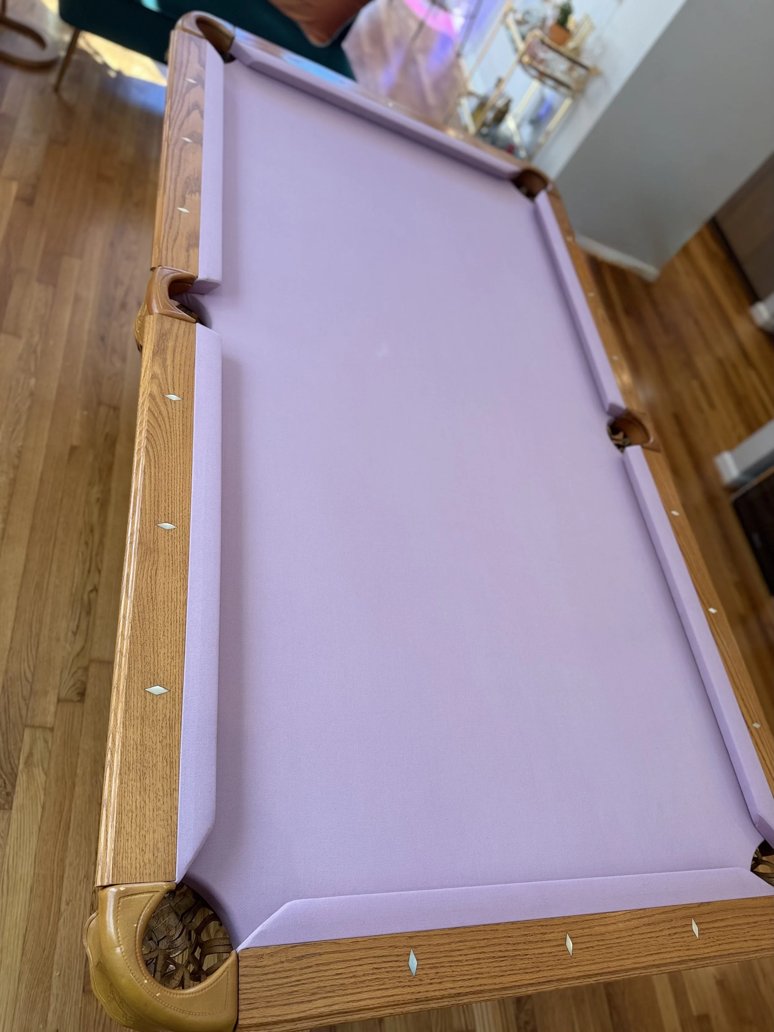 A wooden bed with a lavender-colored mattress and decorative headboard, situated on a hardwood floor in a room with various furniture and artwork in the background.