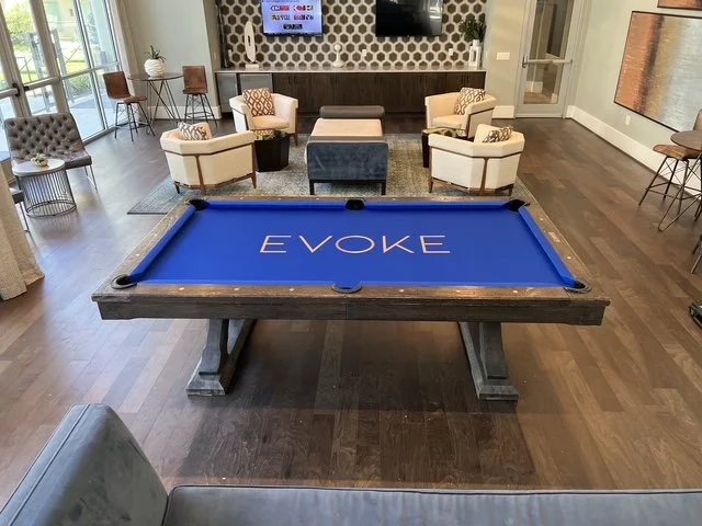 Indoor lounge area with a blue evoking pool table in the center, surrounded by chairs and a TV on the wall.