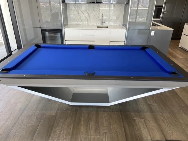 Modern room with a blue pool table with black pockets, white base, and gray borders, surrounded by wooden flooring and kitchen cabinets in the background.